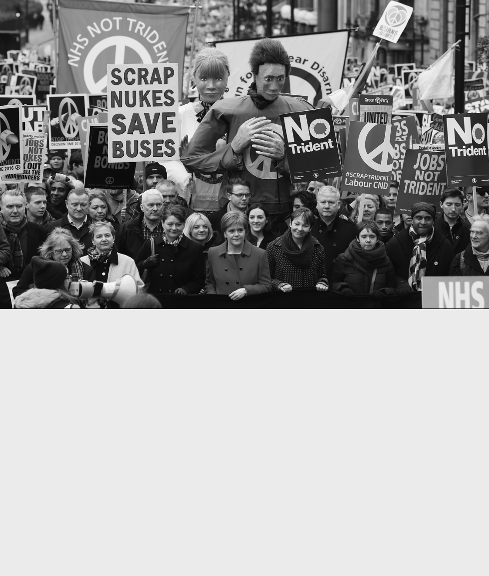 Protest rally with many people holding signs opposing nuclear weapons and supporting the NHS, with some signs reading "SCRAP NUKES SAVE BUSES" and "NO Trident," and large puppets resembling politicians.