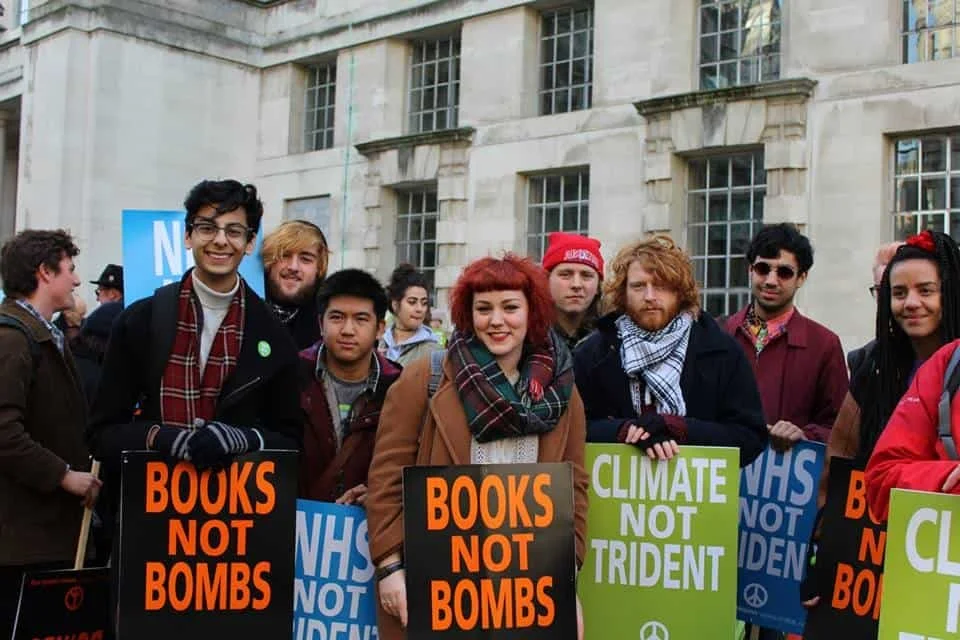Group of young people protesting outdoors, holding signs that say "BOOKS NOT BOMBS" and "CLIMATE NOT TRIDENT" in front of a building.