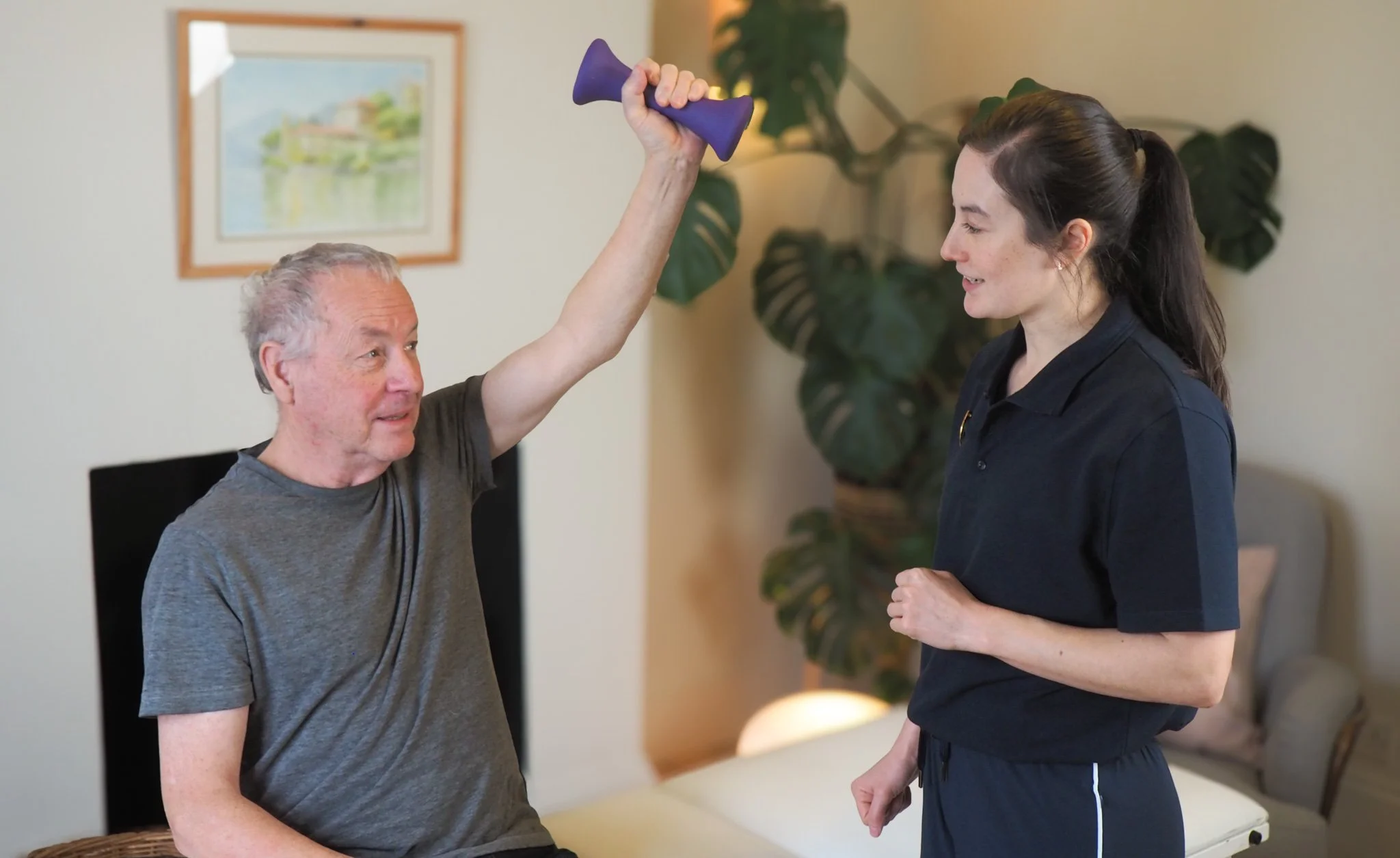 A male client sitting on a treatment couch doing arm exercises with a purple dumbbell while a physiotherapist watches and encourages him.
