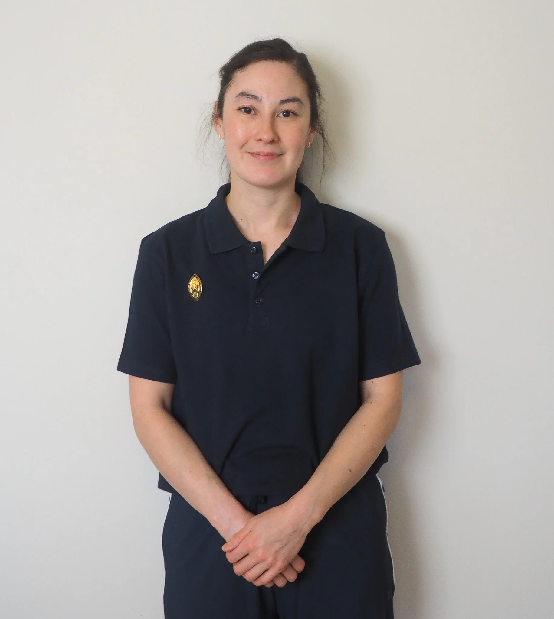 A profile photograph of a physiotherapist - a young woman with dark hair in a ponytail, wearing a navy polo shirt with a logo pin, standing against a plain white wall, smiling with hands clasped in front.
