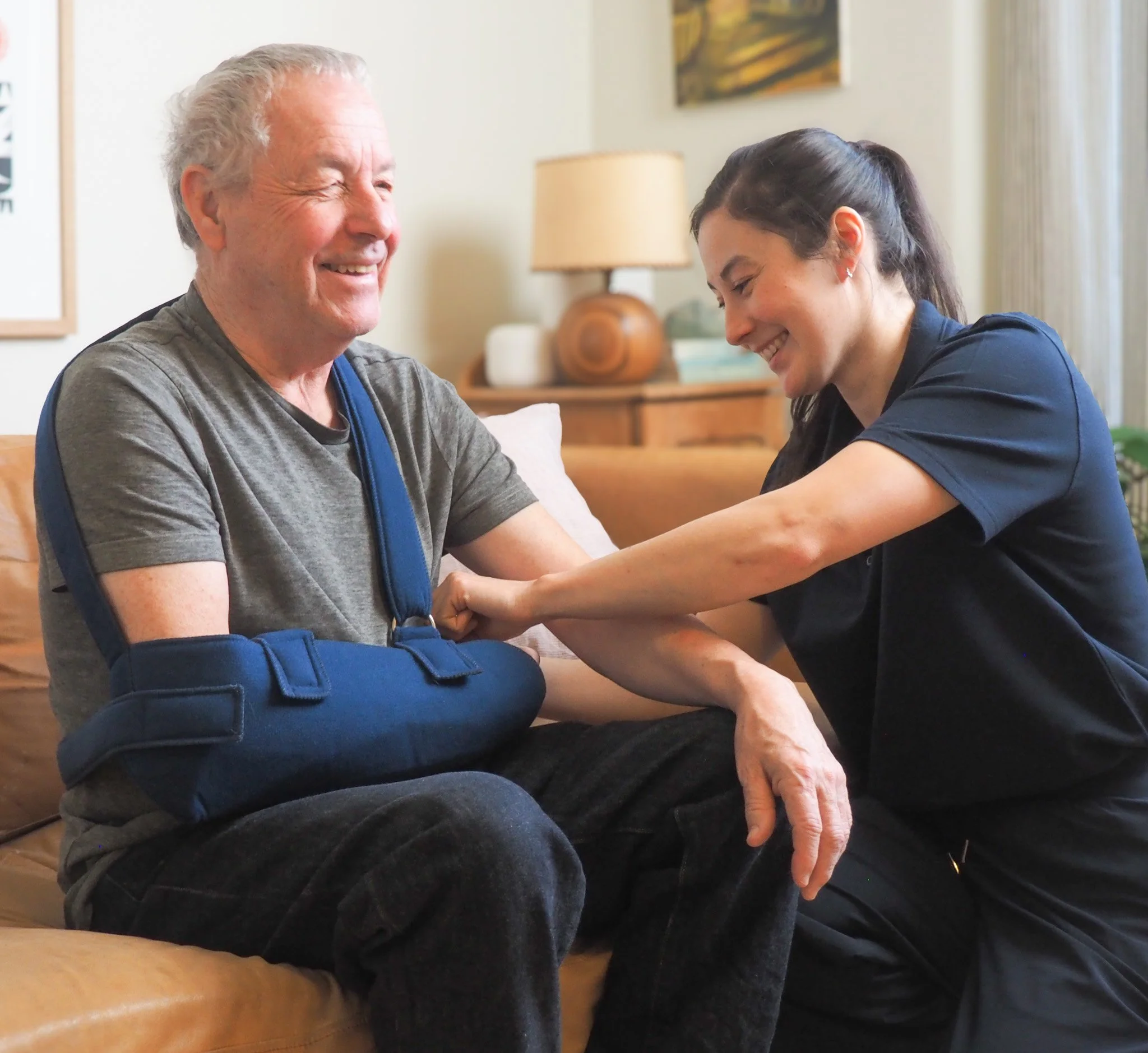 A physiotherapist helps a senior man by adjusting his arm sling and smiling at him inside a cozy living room.