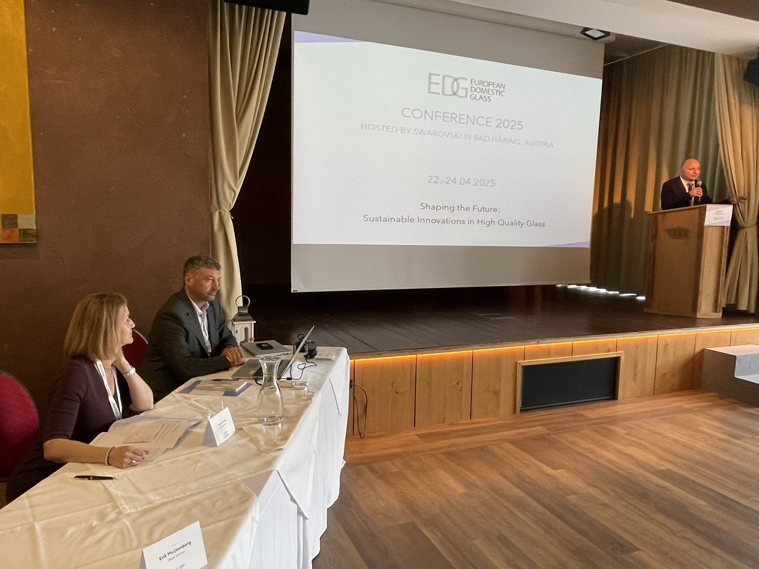 A conference room with a presentation on a large screen showing details of the European Domestic Glass Conference 2025 hosted in Bad Haring, Austria, from April 22 to 24, 2025, focusing on sustainable innovations in high quality glass. There is a spe