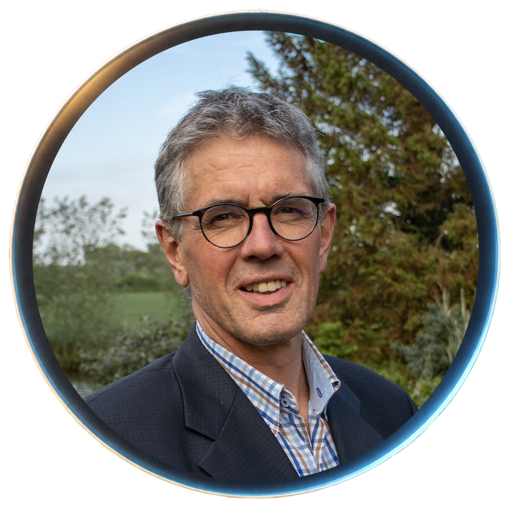 A middle-aged man with gray hair, glasses, wearing a dark blazer and a checkered shirt, standing outdoors in front of trees and a clear sky.
