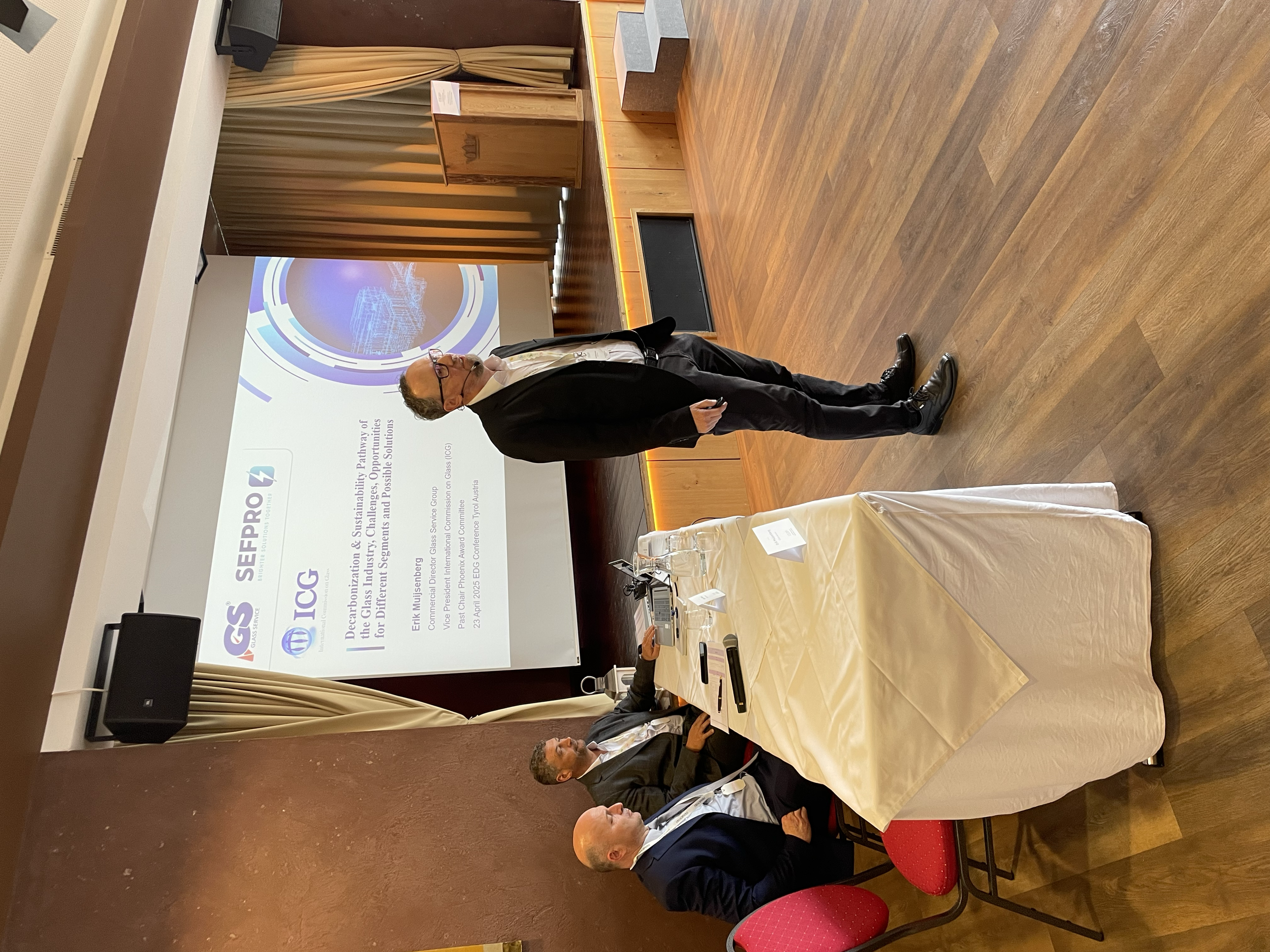 A man in a black suit and glasses giving a presentation at a conference, with a slide about sustainability, on a screen behind him. Three men are seated at a table watching the presentation, with phones and notes in front of them.
