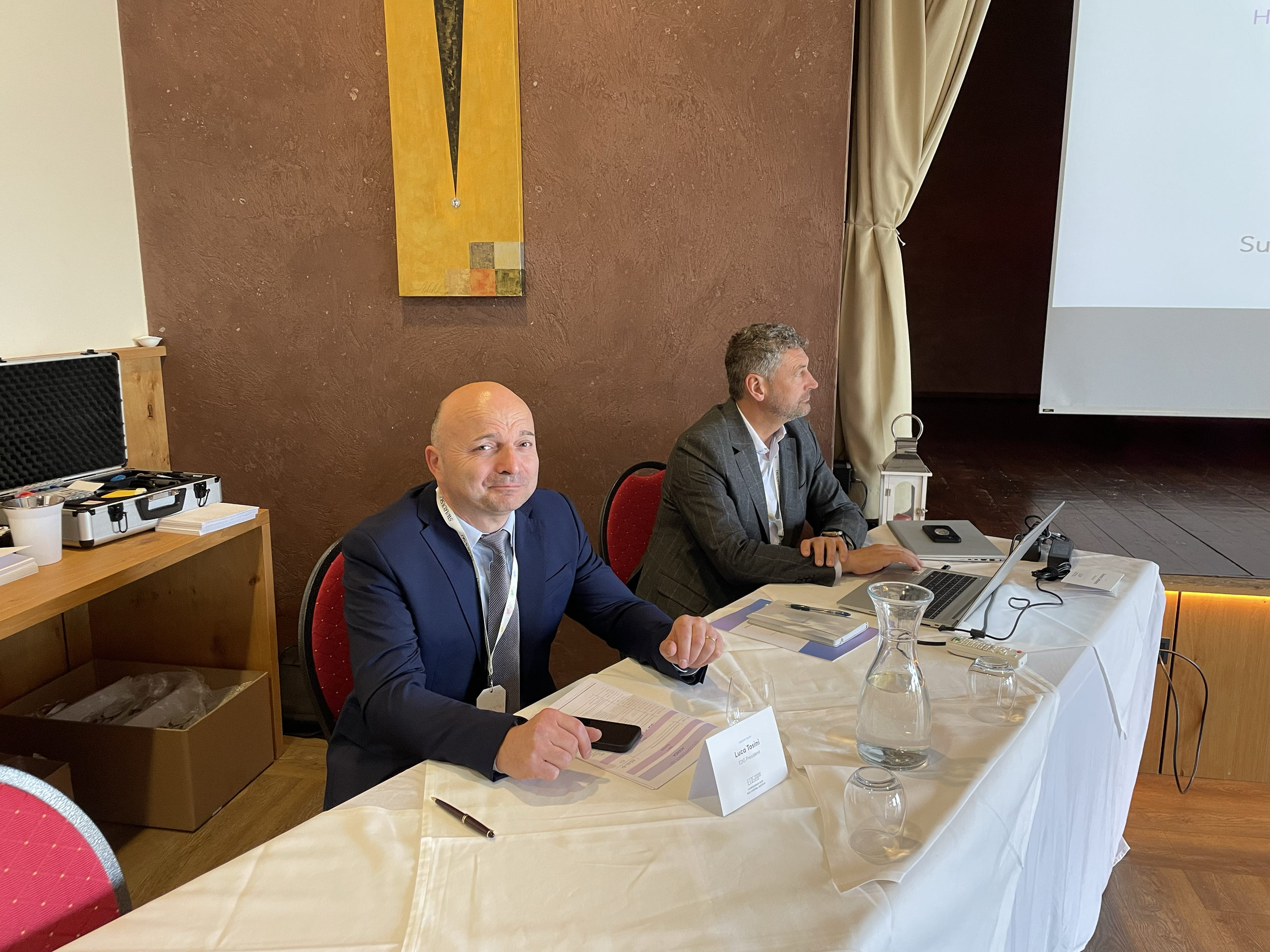 Two men in suits sitting at a conference table in a meeting room with presentation screen to the right. One man faces forward with a slight smile, and the other is looking to the side. The table has laptops, papers, a glass pitcher of water, and name