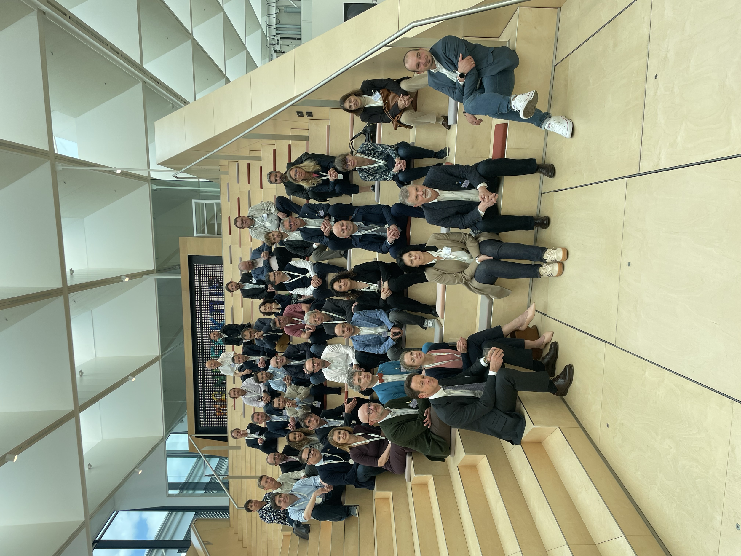 Group of people gathered in a modern indoor space with wooden stairs and large windows. Some individuals sit on the stairs, while others stand around or pose for the photo. The setting appears to be a professional or conference environment.