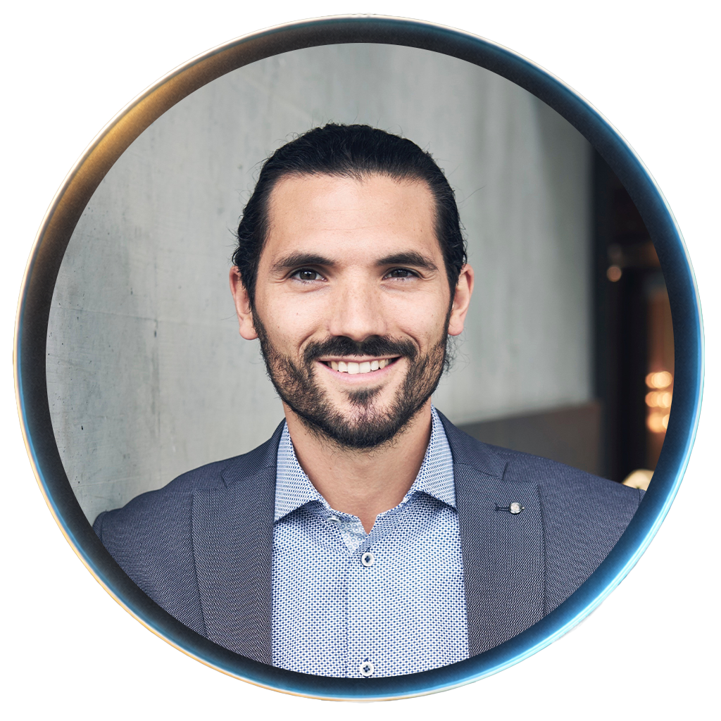 A smiling man with dark hair, a beard, wearing a suit and a patterned dress shirt, looking directly at the camera.
