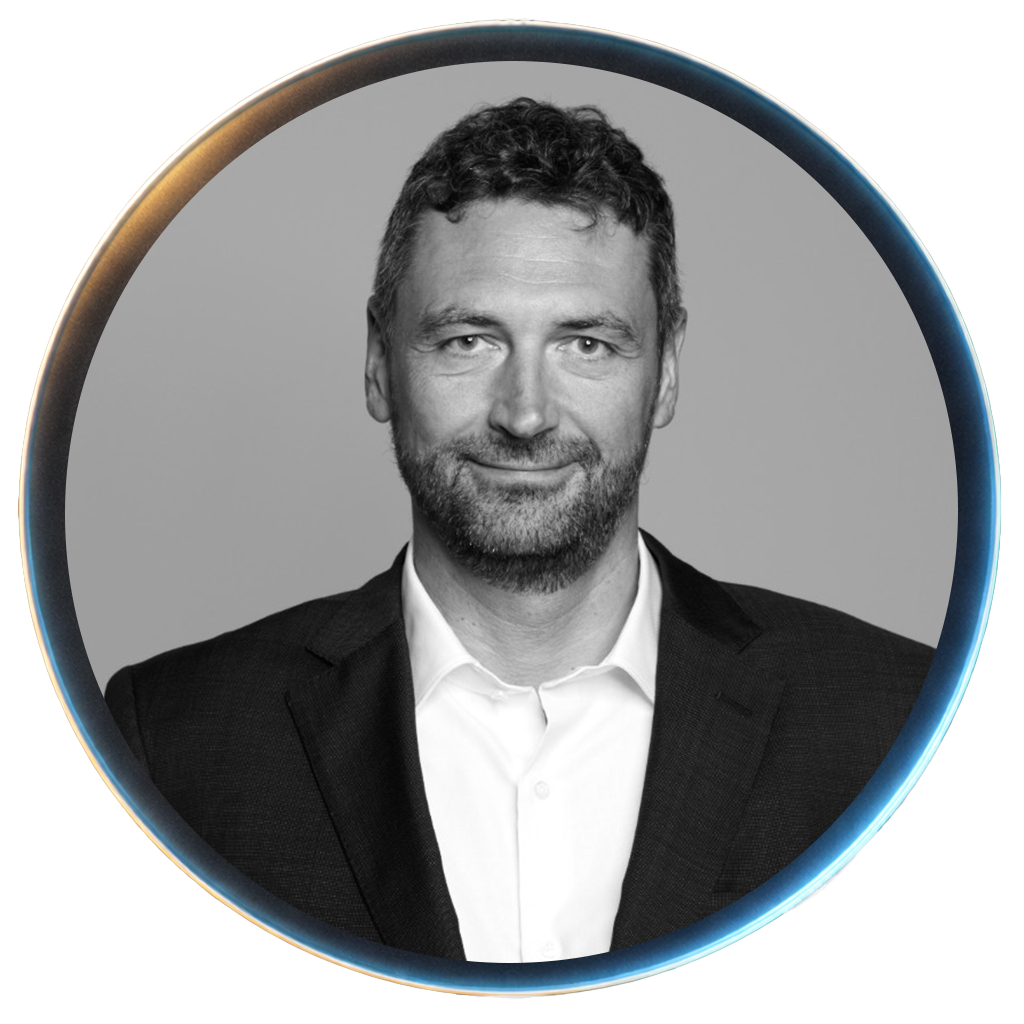 Black and white professional portrait of a man with curly hair and a beard, wearing a suit and white shirt, smiling slightly.