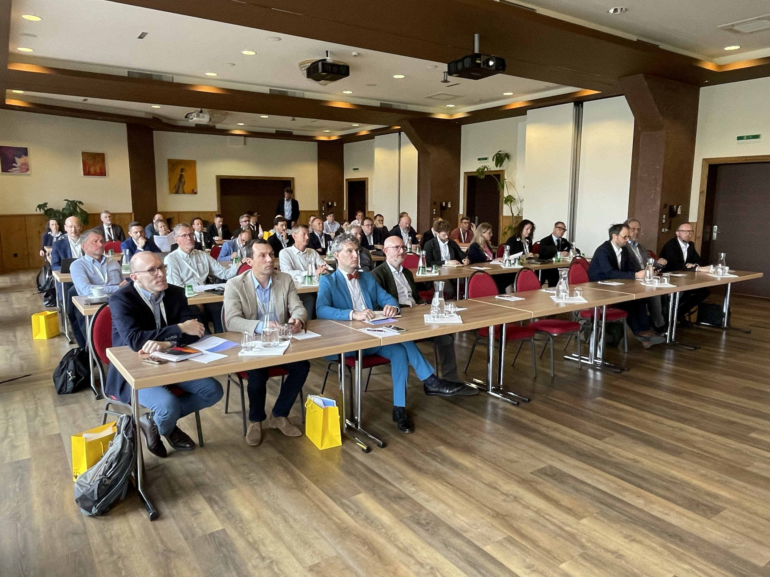 A conference room filled with professionally dressed attendees sitting at tables, listening to a speaker or presentation. The room has wood flooring, modern decor, and wall art.