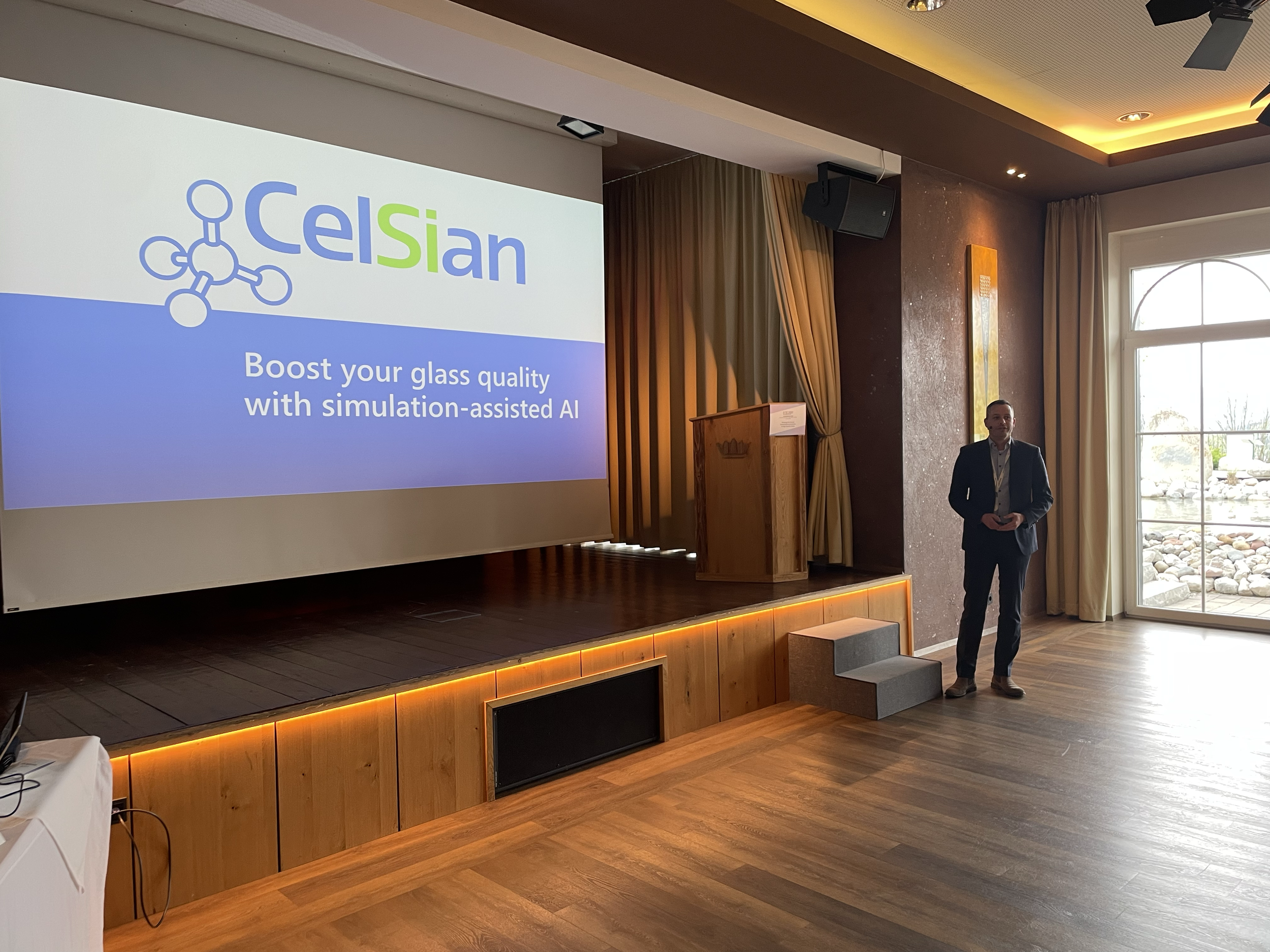 A man in a dark suit giving a presentation in a room with wooden flooring, standing next to a large screen displaying the logo and slogan of Celsian, which offers simulation-assisted AI to boost glass quality. There are curtains, a window showing roc