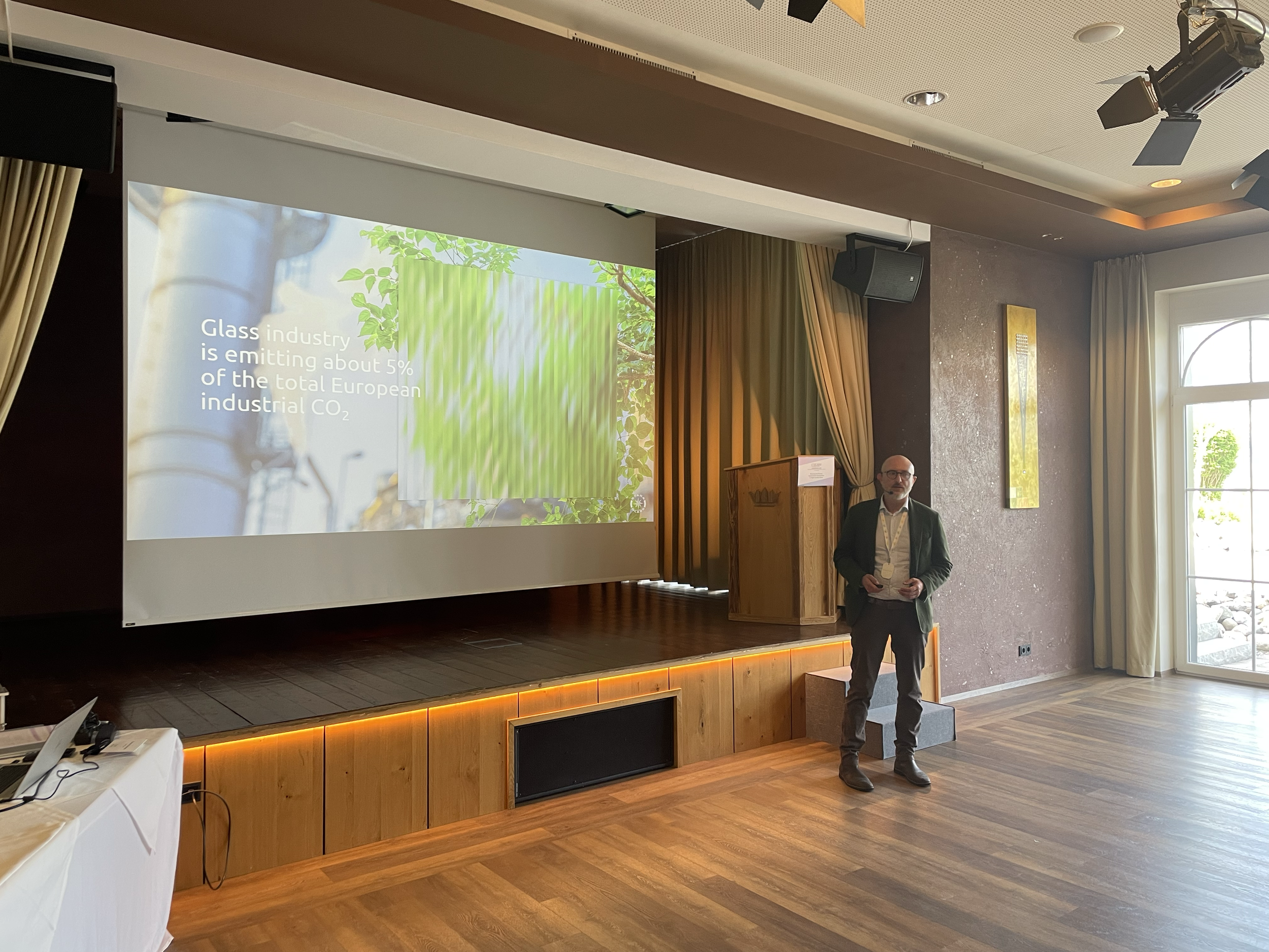 A man standing on a small platform in front of a large screen in a conference room, giving a presentation about the glass industry's CO2 emissions. The screen displays a slide with text and images related to the topic, with a podium nearby and natura