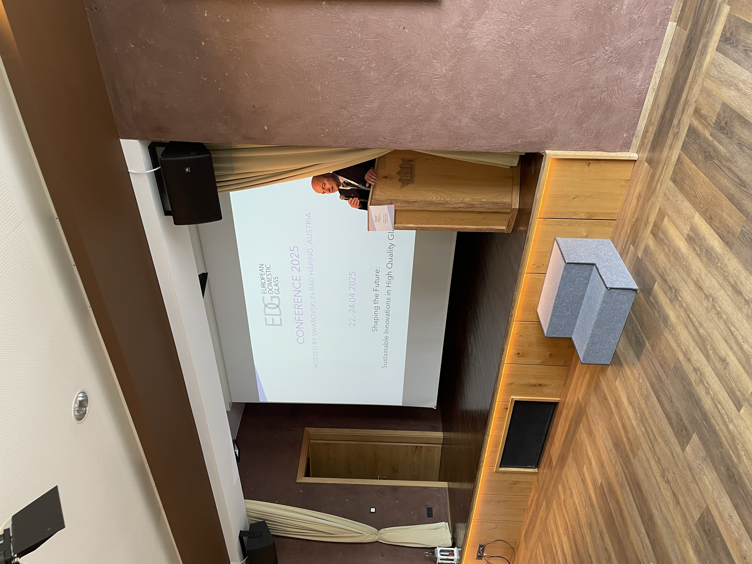 A man in a suit giving a presentation at a conference with a large screen behind him displaying the conference details and date.
