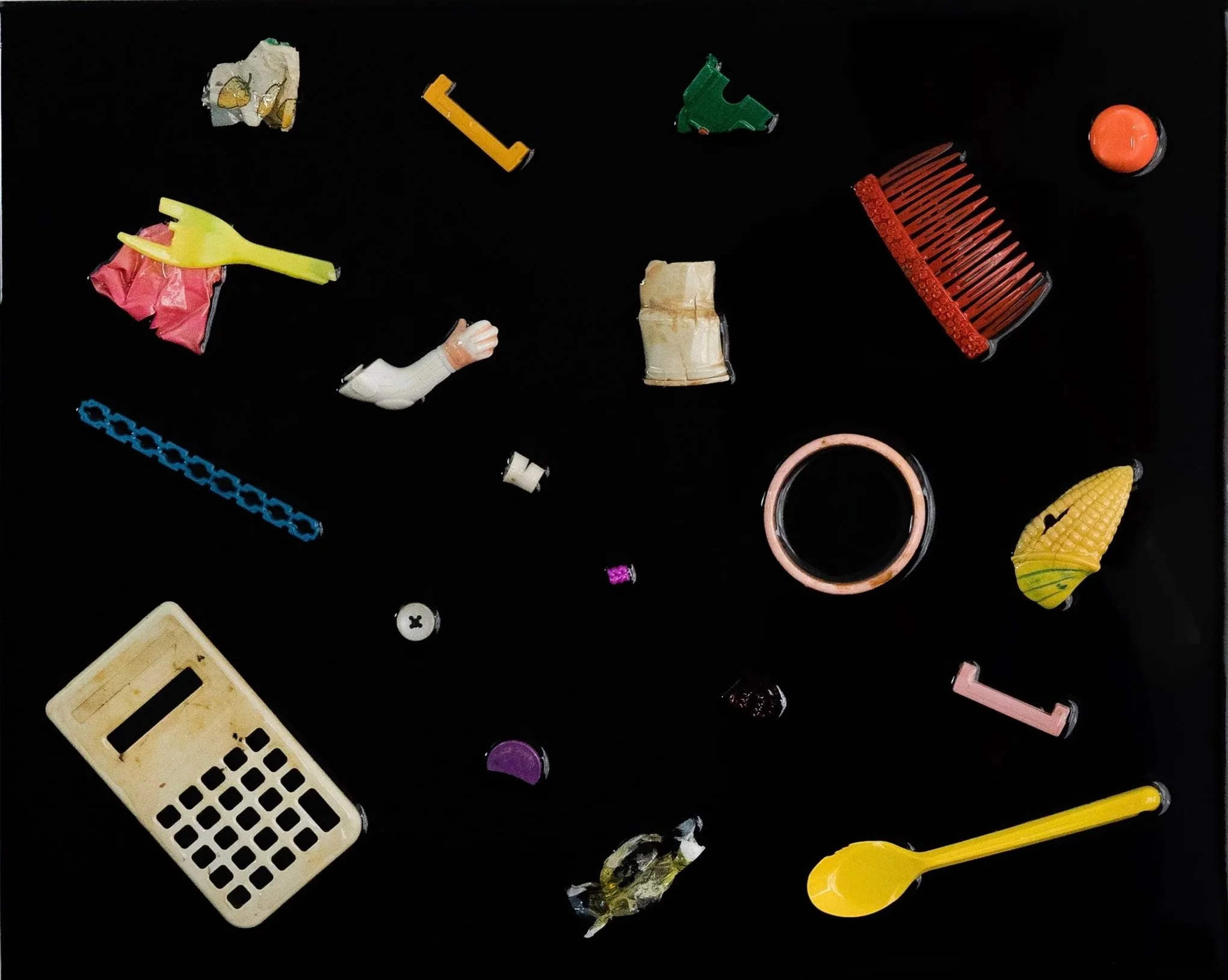 Various small plastic and rubber objects on a black background, including a yellow fork, a red comb, a peach circle, a small white basket, a purple piece, and a gift-wrapped item.