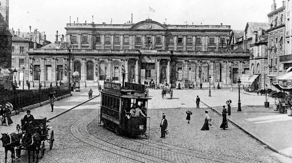 Une photo en noir et blanc d'une place urbaine avec un bâtiment emblématique en arrière-plan, une tramway en mouvement, des personnes marchant, des chevaux avec une calèche, et des lampadaires de style ancien.