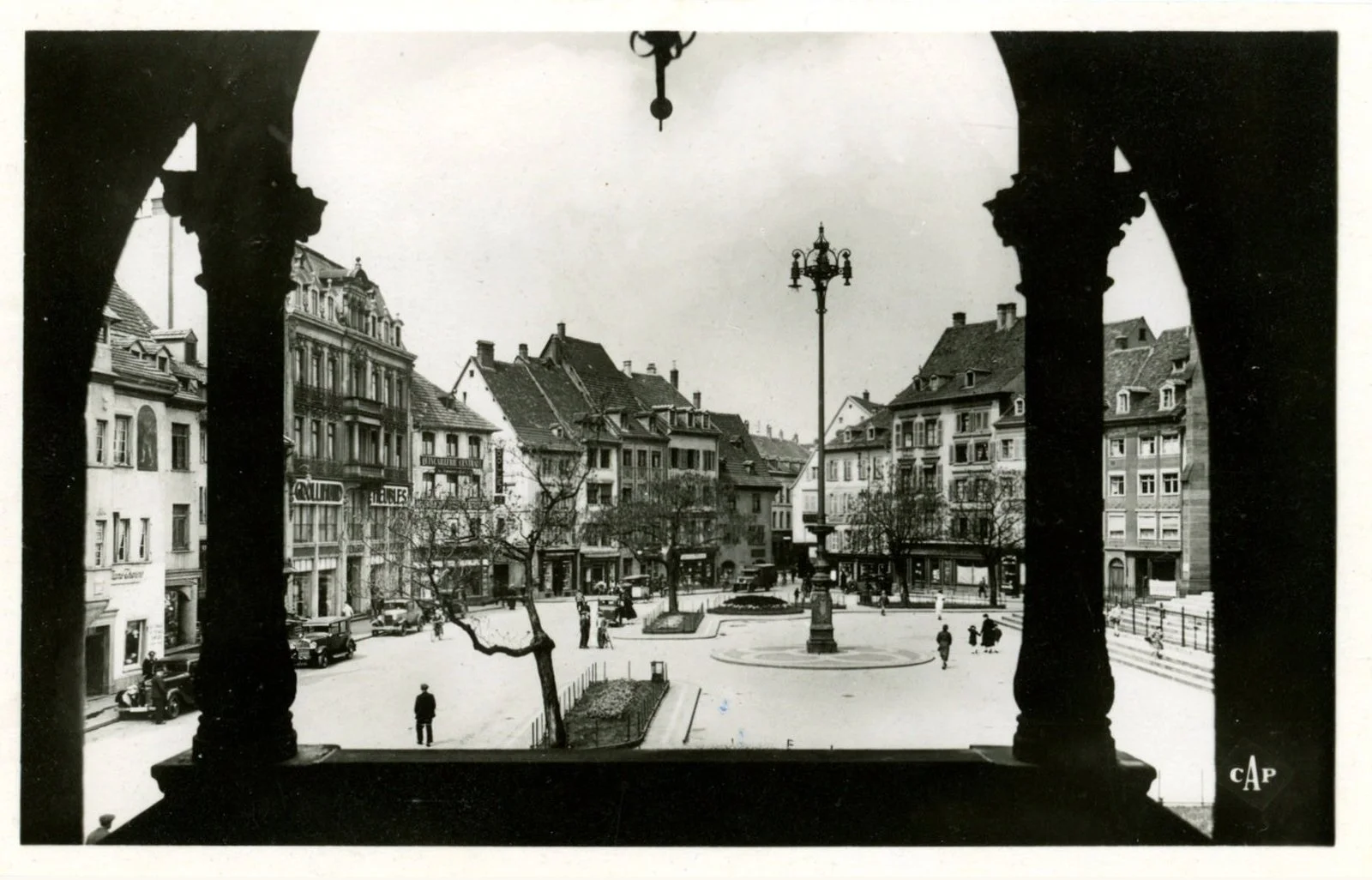 Vue en noir et blanc d'une place urbaine depuis une fenêtre, avec des bâtiments anciens, des arbres dénudés, une fontaine centrale et des lampadaires, avec quelques personnes et voitures.