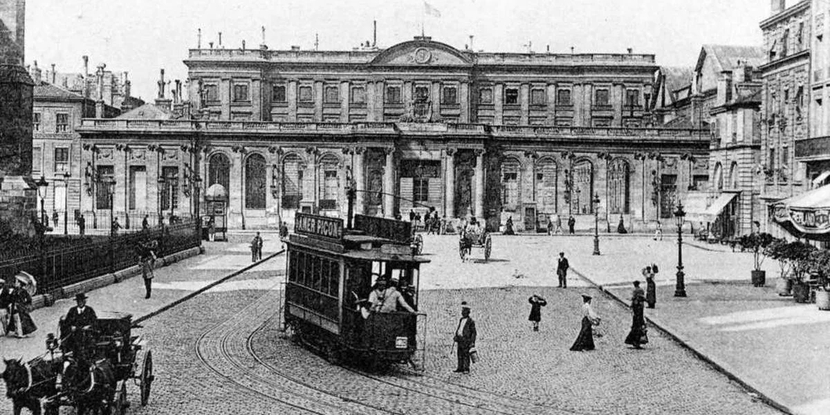 Une image en noir et blanc représentant une place urbaine historique avec un bâtiment architectural en arrière-plan, un tramway en mouvement, des personnes marchant et des calèches.