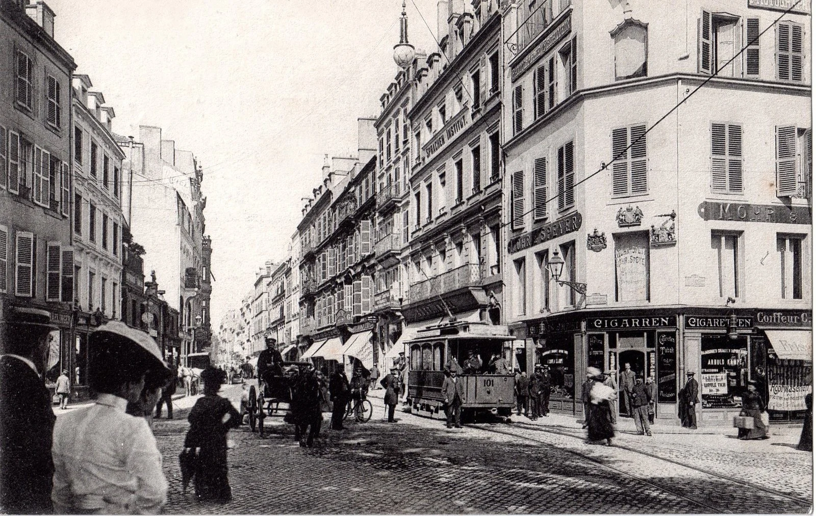 Une rue animée datant probablement du début du 20e siècle avec un tramway, des gens, des boutiques et des bâtiments anciens.