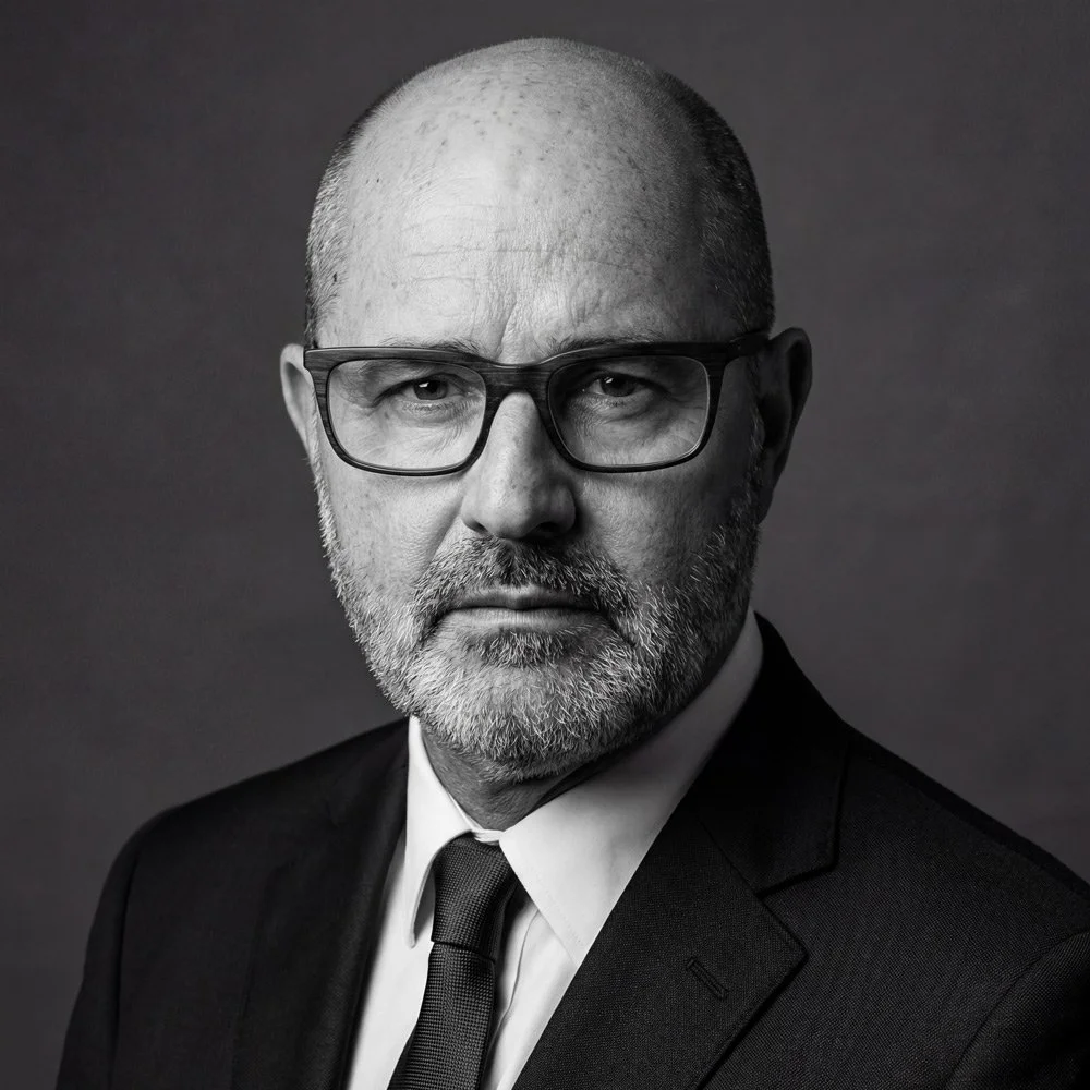 Portrait en noir et blanc d'un homme barbu portant des lunettes, un costume et une cravate, regardant sérieusement vers l'objectif.