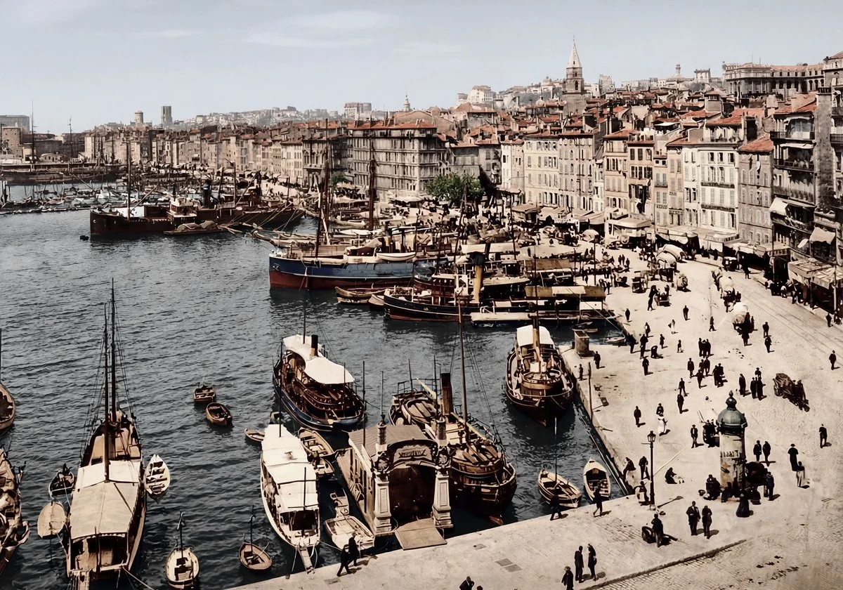 Vue d'un port avec des bateaux amarrés et une promenade animée bordée de bâtiments anciens, avec une église pointue en arrière-plan, sous un ciel clair.