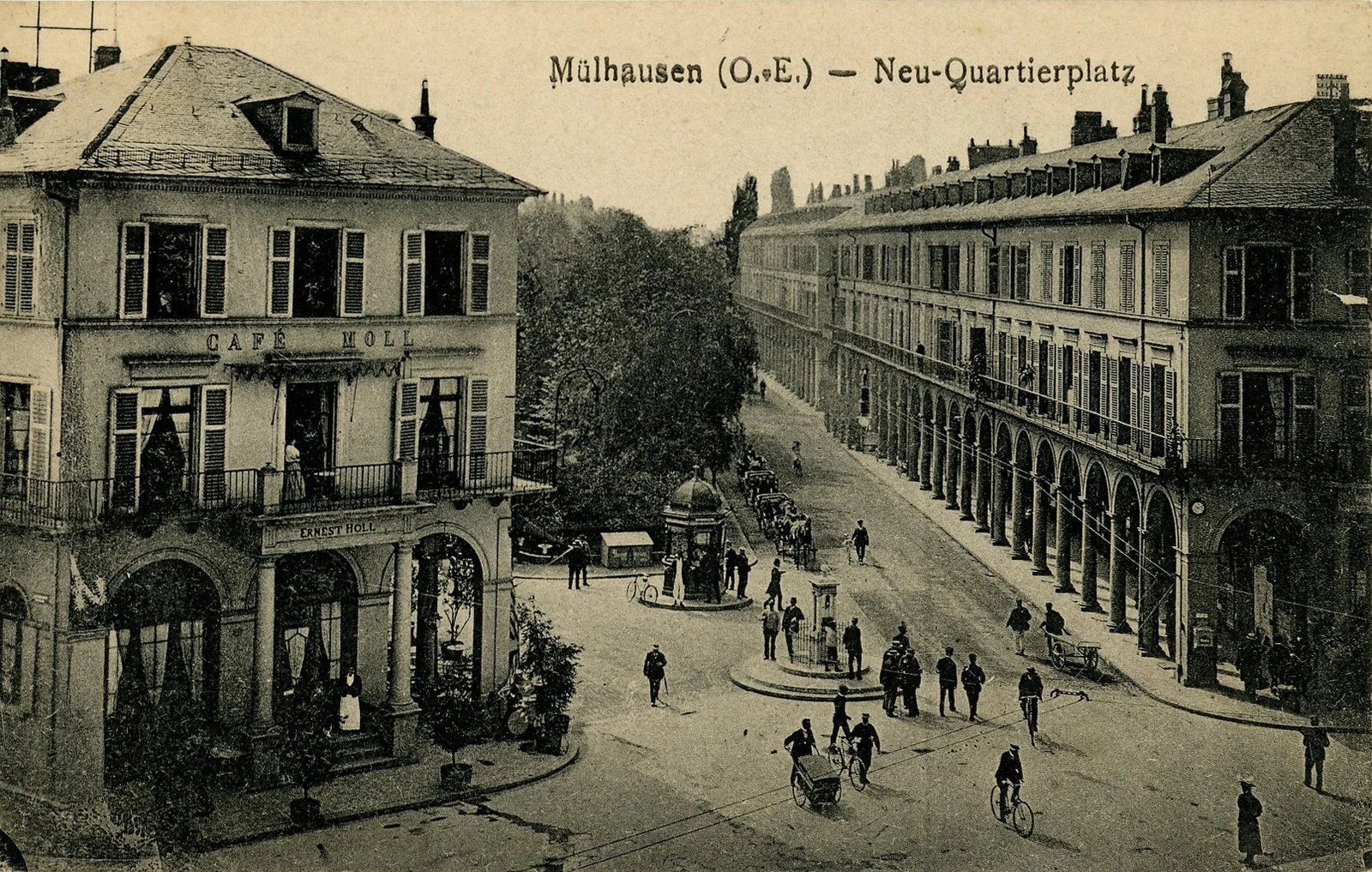 Une vieille photo en noir et blanc montre une rue animée à Mülhausen avec des piétons, des cyclistes et des voitures anciennes. Sur la gauche, un bâtiment avec une enseigne "Café Möll" et une terrasse, sur la droite un long immeuble avec une arcade au rez-de-chaussée. Au centre, une fontaine ou un kiosque en pierre, entouré de quelques personnes.