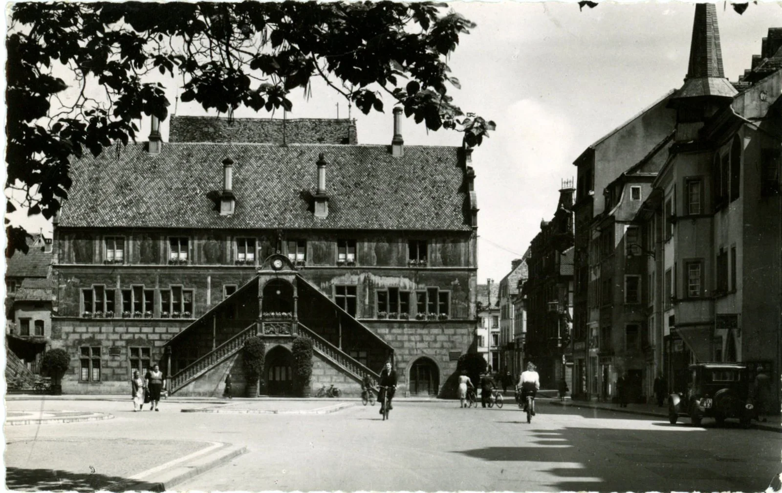 Une place urbaine avec des personnes qui font du vélo, une voiture ancienne et un bâtiment historique avec un escalier extérieur, surement dans une ville européenne.