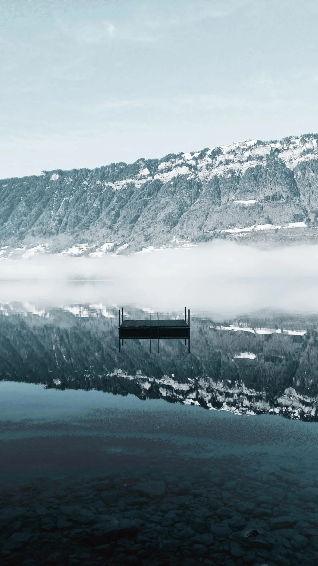 Ein ruhiger See im Winter, umgeben von schneebedeckten Bergen, mit einer kleinen Plattform im Wasser und einer klaren, spiegelglatten Wasseroberfläche, die die Berge und Plattform reflektiert.