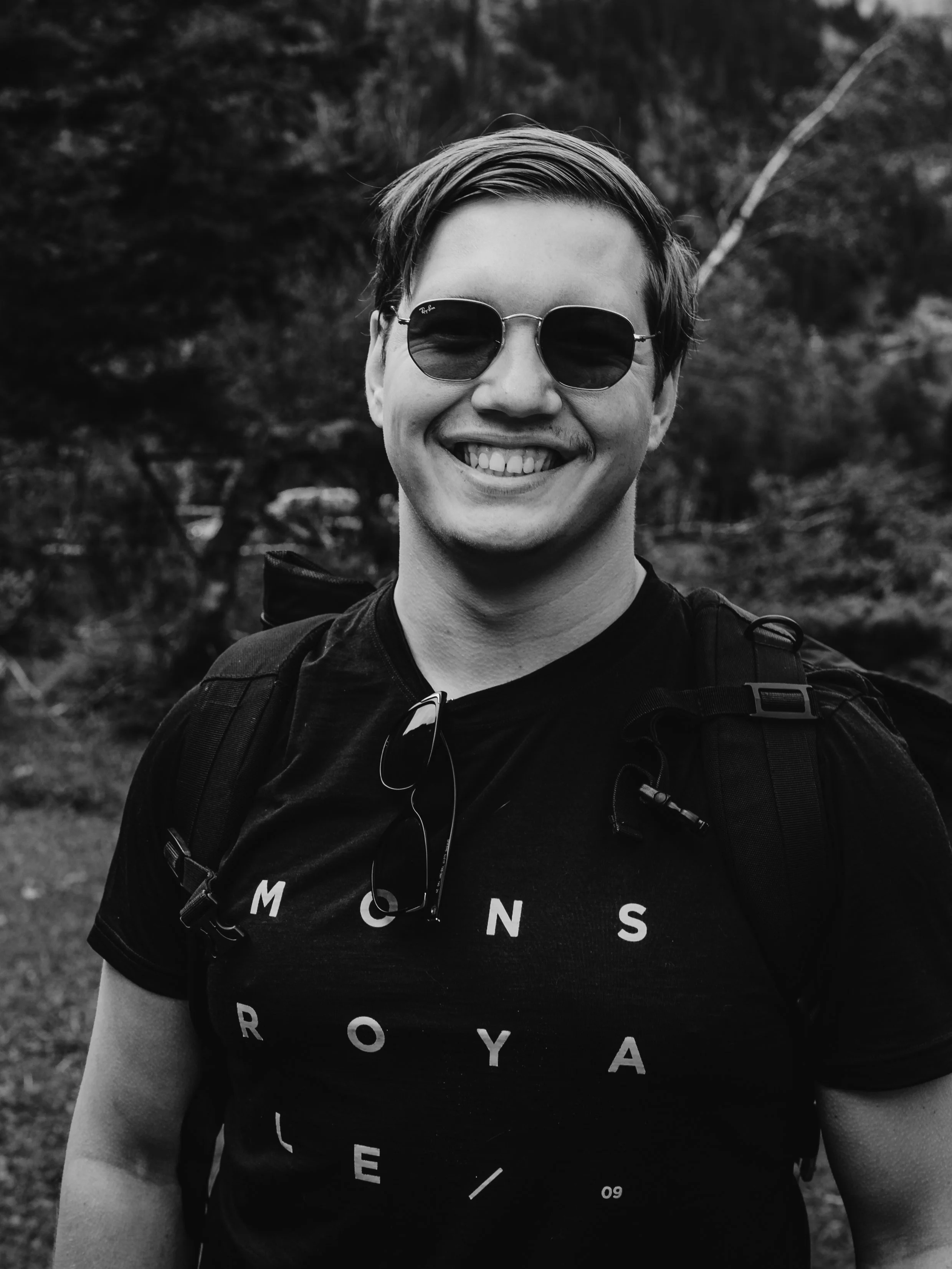 Ein lachender junger Mann trägt Sonnenbrille, T-Shirt und Rucksack, steht im Freien in einer natürlichen Umgebung, wahrscheinlich im Park oder Wald.