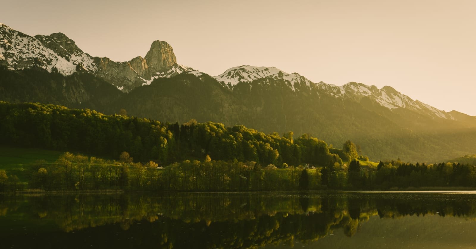 Berglandschaft mit schneebedeckten Gipfeln, grünen Wäldern und einem See im Vordergrund bei Sonnenuntergang.