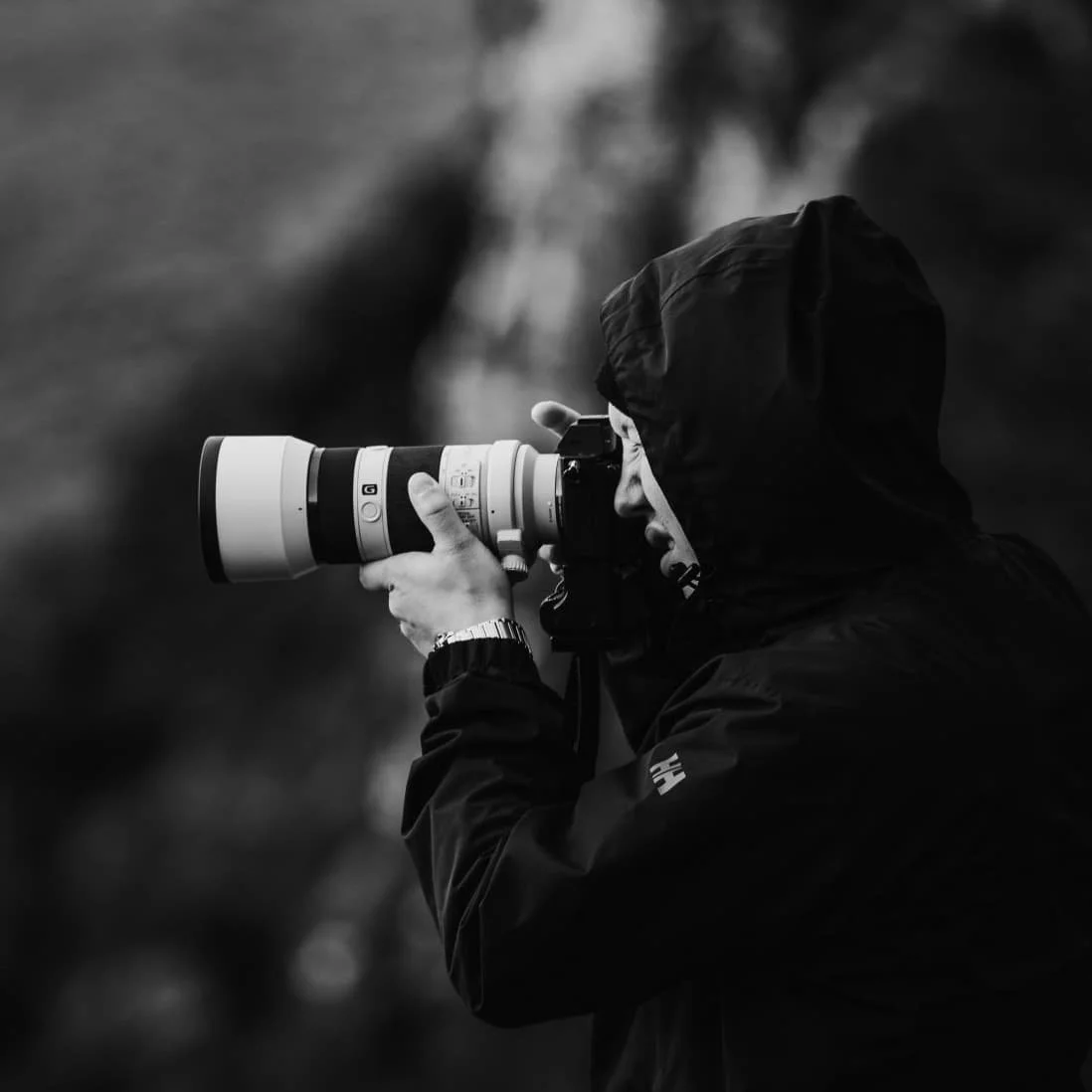 Person mit dunkler Regenjacke und Kapuze fotografiert mit professioneller Kamera mit Teleobjektiv, im Freien bei bewölktem Himmel, Schwarz-Weiß-Fotografie.