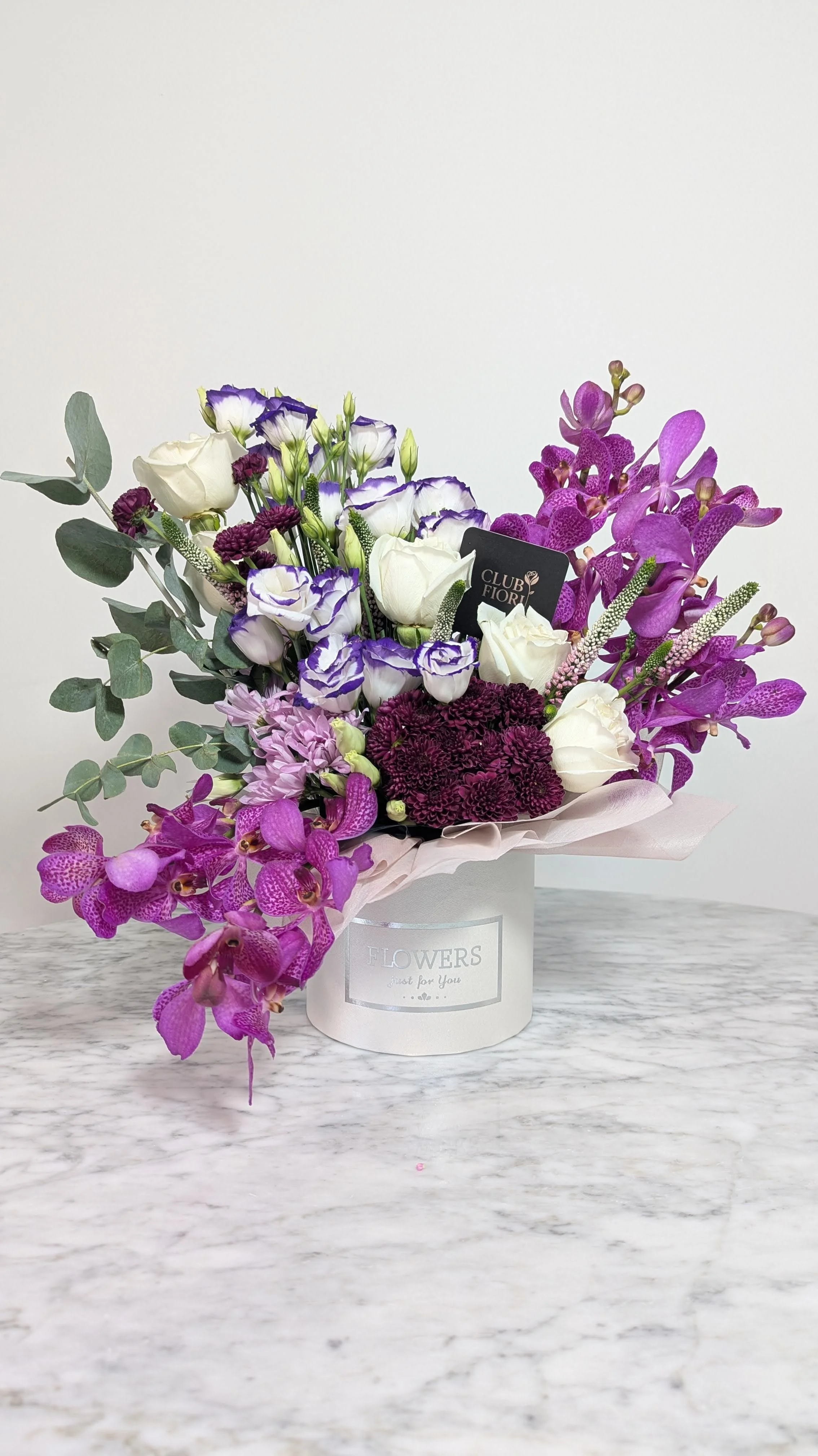 A bouquet of purple, white, and green flowers in a white box with the label 'Flowers just for You' on a marble surface.
