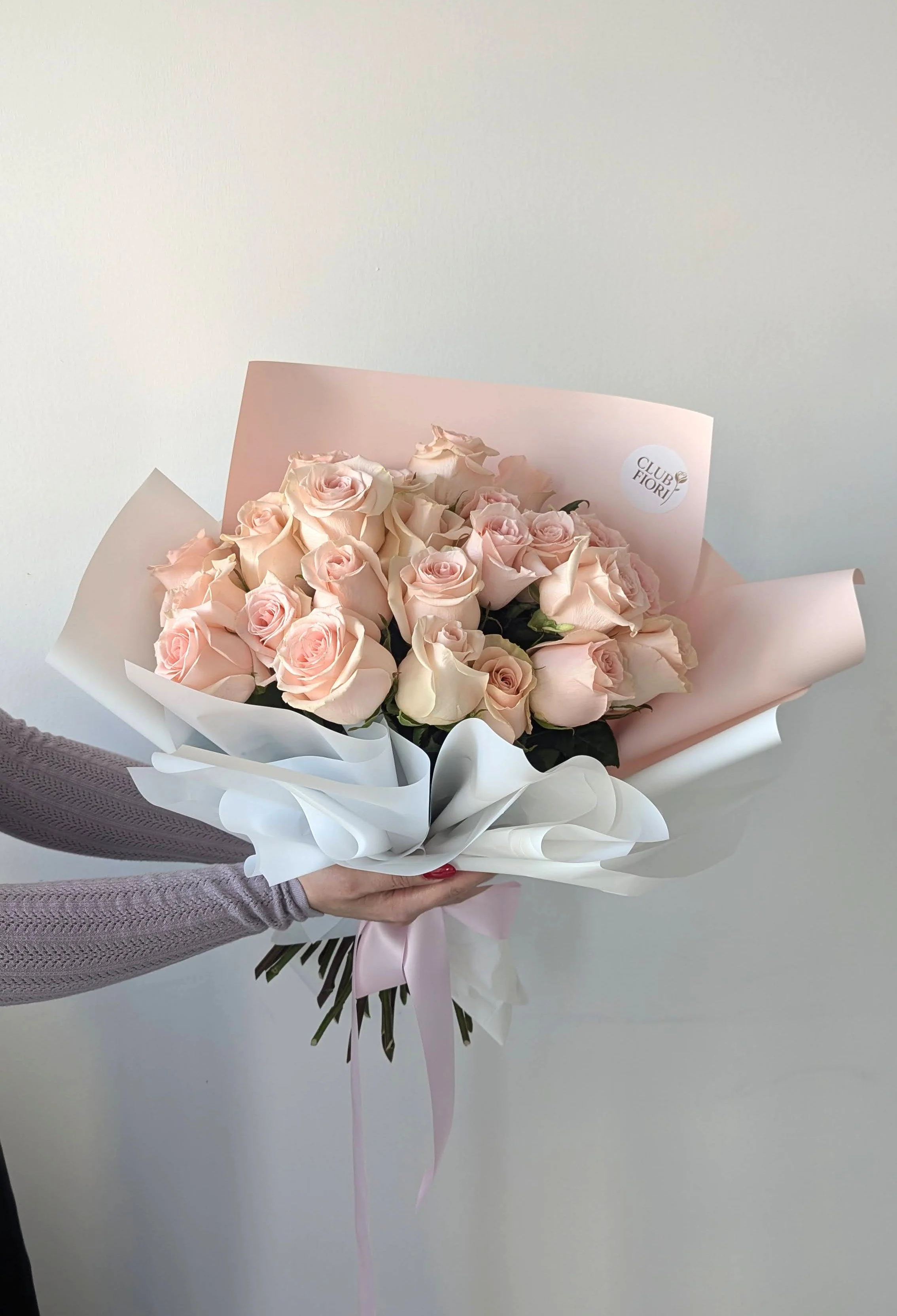 A person holding a bouquet of light pink and peach roses wrapped in pink, white, and gray paper.