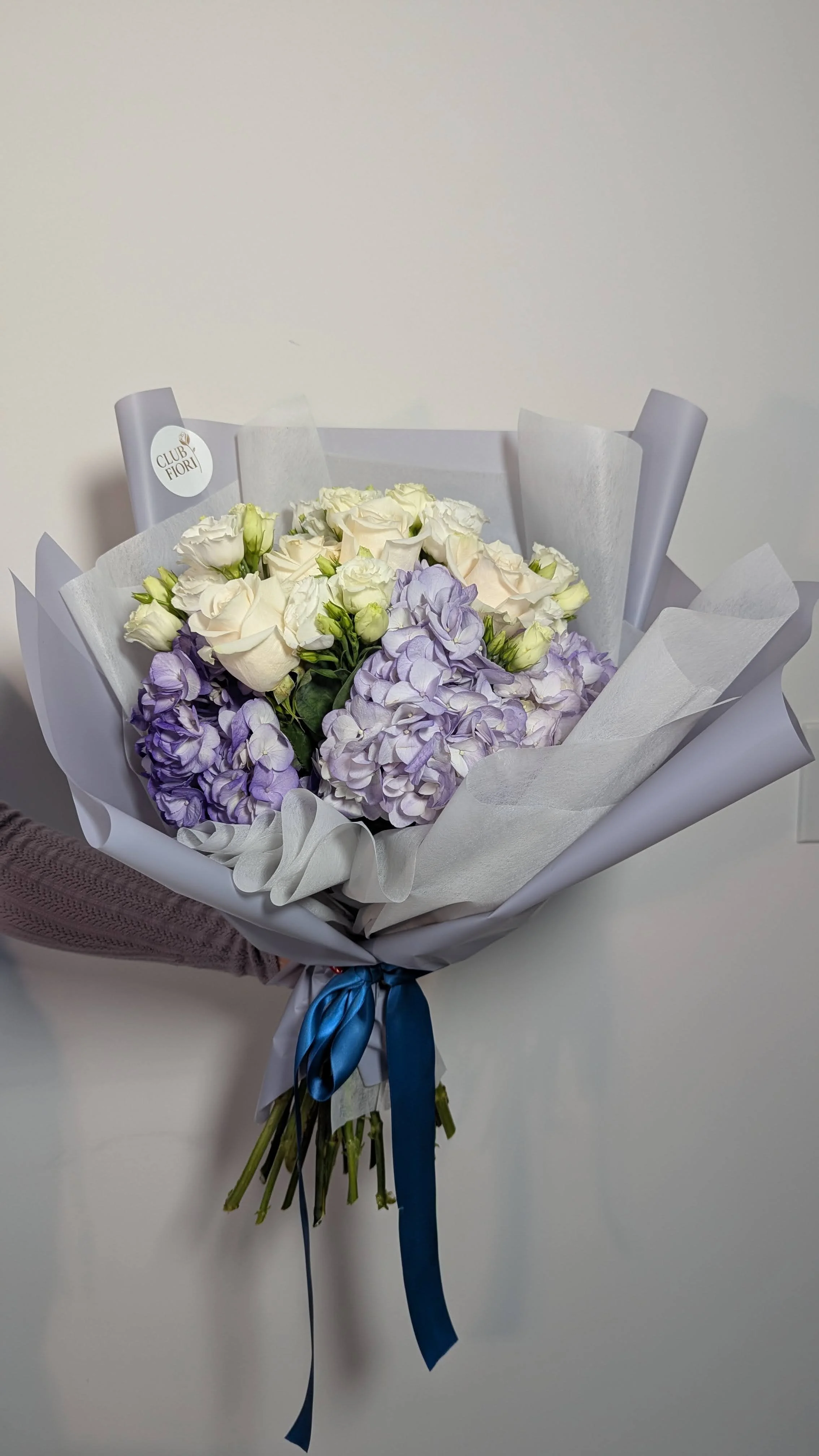 A bouquet of white roses and light purple hydrangeas wrapped in white paper with a blue ribbon.