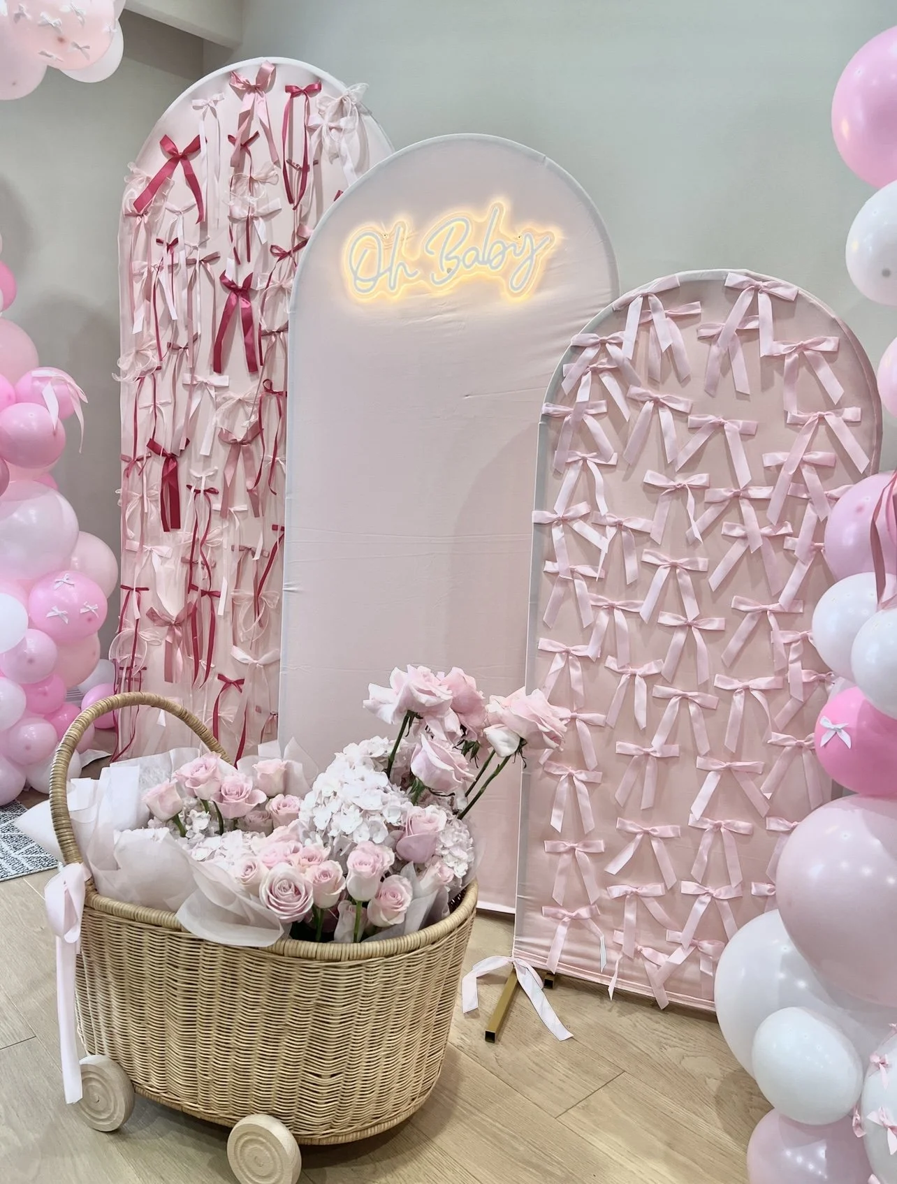 Decorative display with pink and white balloons, pink ribbons on fabric backdrops, a neon sign that says 'Oh Baby', and a basket of pink and white flowers.