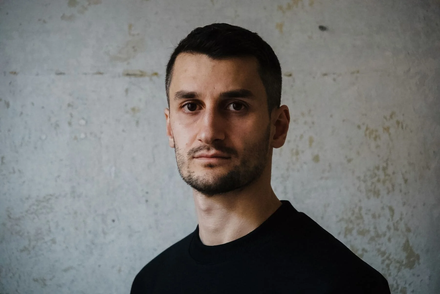 Ein junger Mann mit dunklem Haar und Bart trägt ein schwarzes Shirt vor einer hellen, leicht strukturierten Wand.
