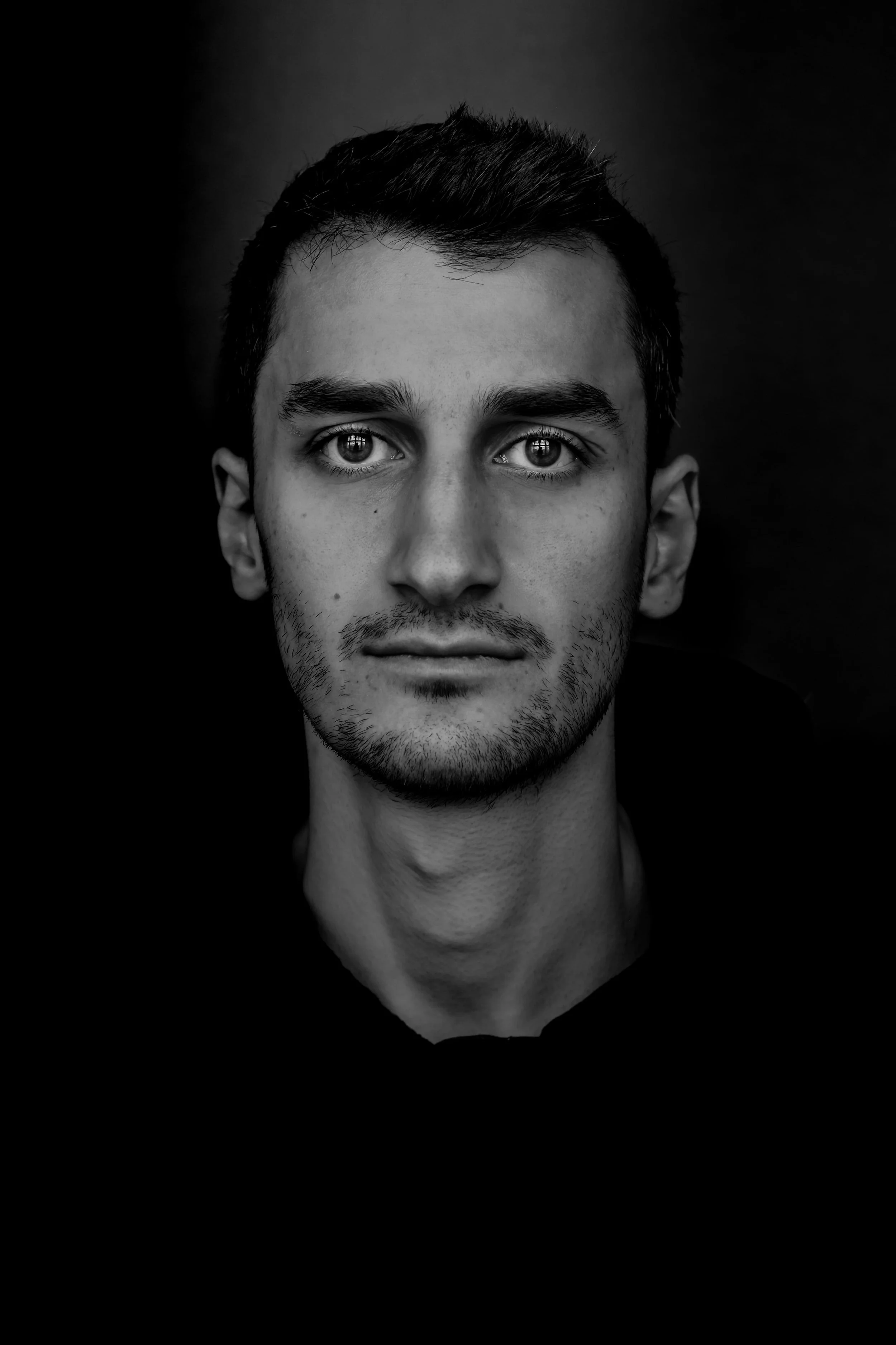 Schwarz-weiß-Porträt eines jungen Mannes mit kurzem dunklem Haar und leichtem Bart, Blick direkt zur Kamera, dunkler Hintergrund.