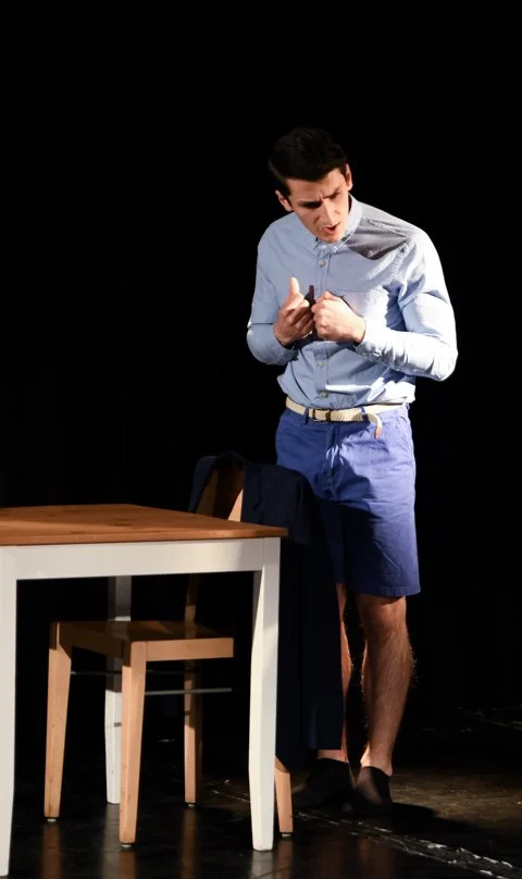 Mann steht an einem Tisch, trägt Hemd und Shorts, wirkt schockiert oder ärgerlich, gegen einen schwarzen Hintergrund.