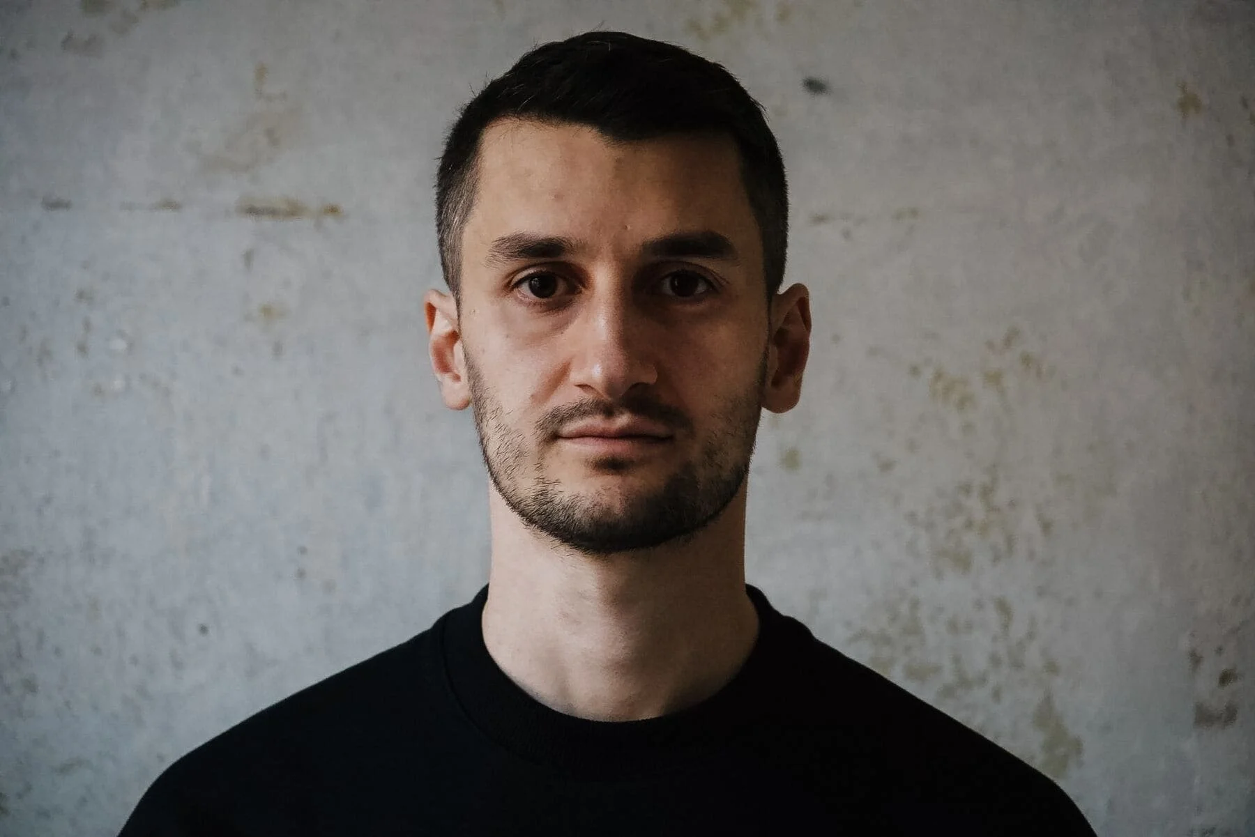 Porträt eines Mannes mit dunklem Haar, braunen Augen und dunklem Hemd vor einer grauen Wand.