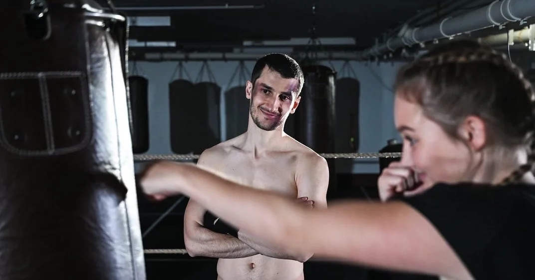 Ein Mann und eine Frau trainieren im Fitnessstudio; die Frau schlägt in den Boxsack, der Mann beobachtet sie lächelnd.