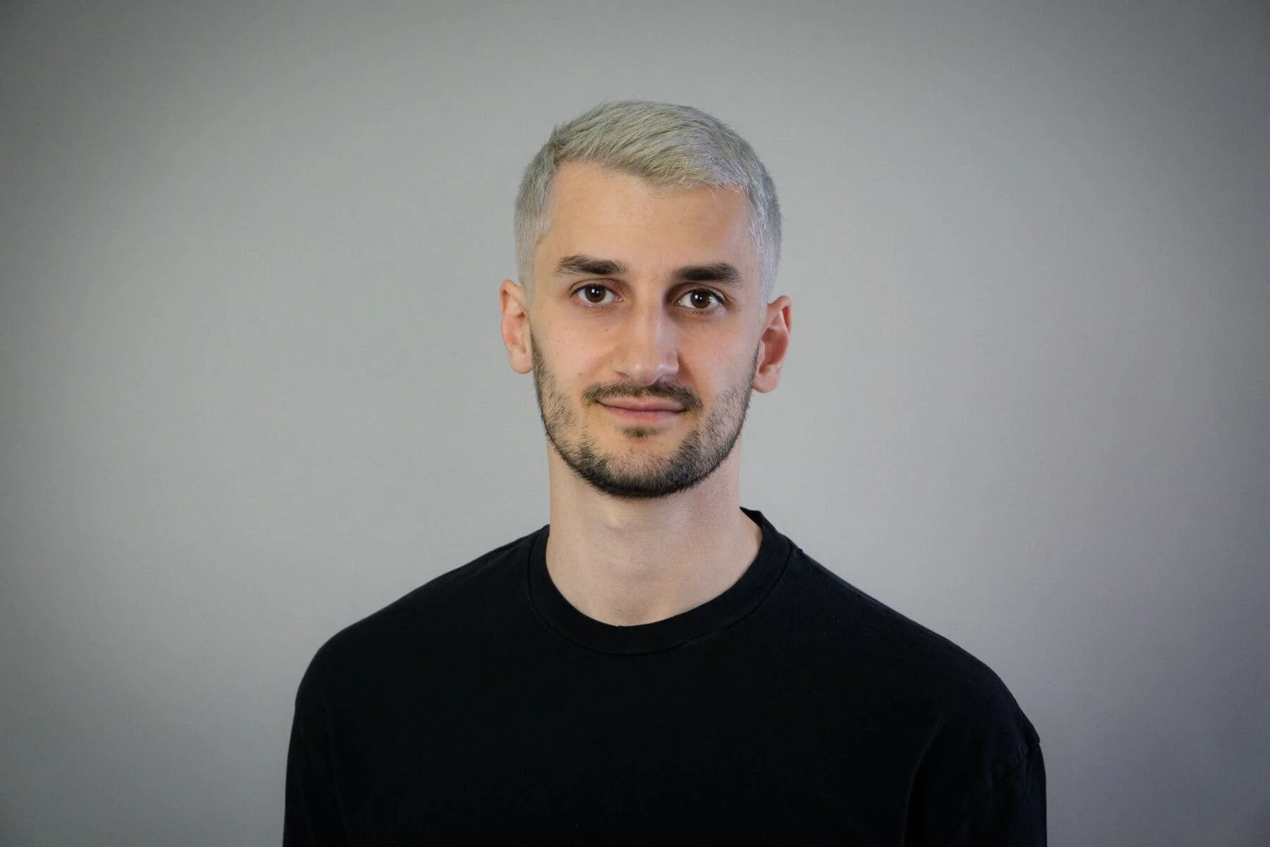 Ein junger Mann mit kurzen grauen Haaren und Bart trägt ein schwarzes T-Shirt, steht vor einem grauen Hintergrund und lächelt leicht.