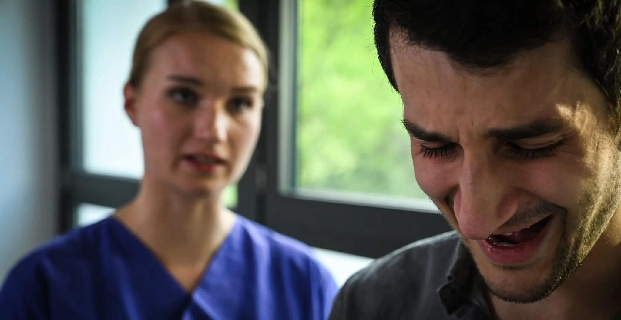 Zwei Menschen stehen nahe beieinander vor einem Fenster, die Frau trägt blauer Kittel und betrachtet den Mann, der lachend nach unten schaut.