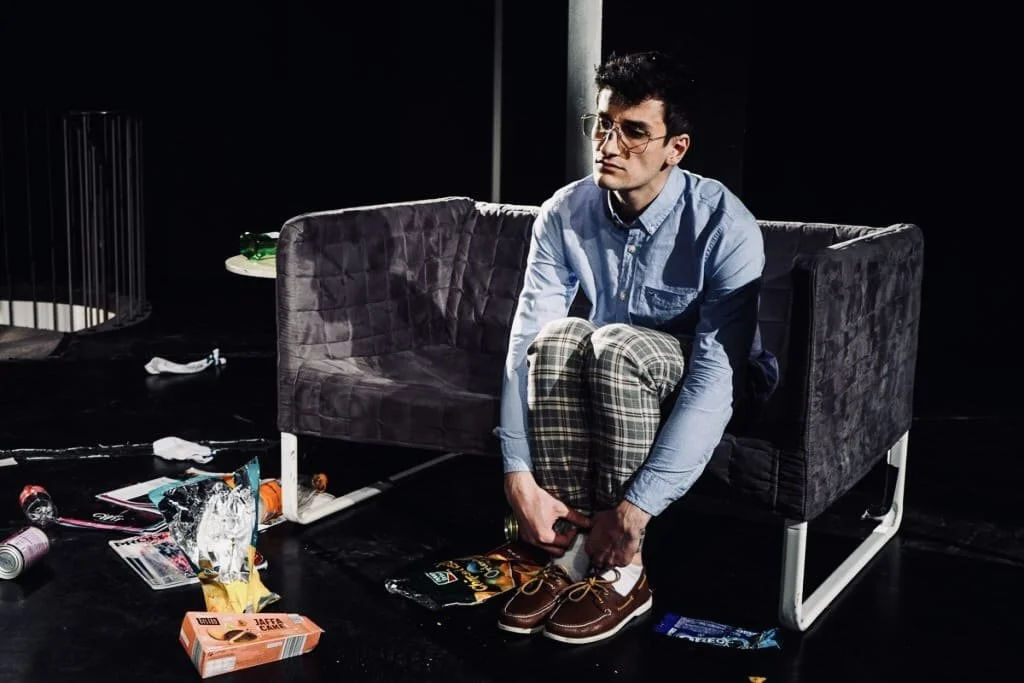 Ein junger Mann sitzt auf einem schwarzen Sofa, wirkt nachdenklich, sammelt seine Schuhe auf, um sie anzuziehen, umgeben von Müll und leeren Verpackungen auf dem Boden, in einem dunklen Raum.