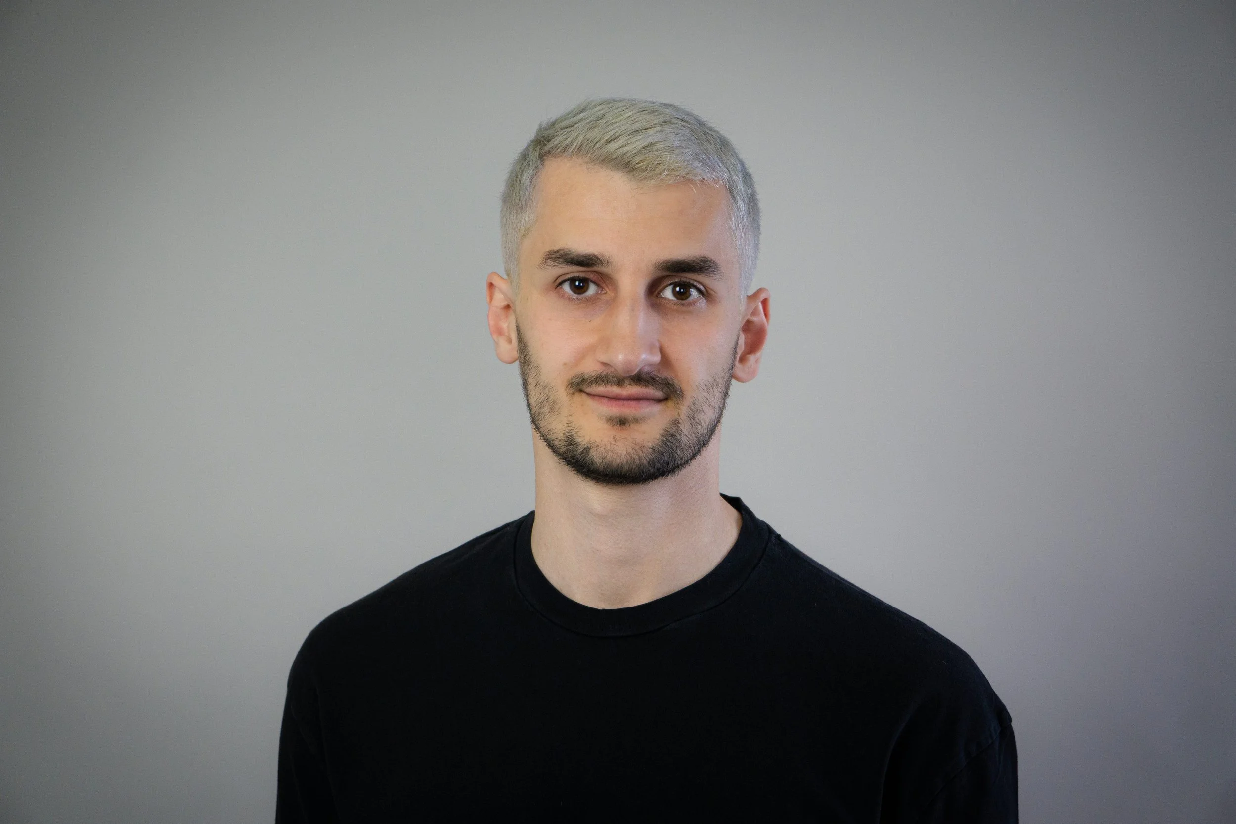 Porträt eines jungen Mannes mit blondem Haar, Kurzhaarschnitt, dunklem Bart, schwarzem T-Shirt vor grauem Hintergrund.