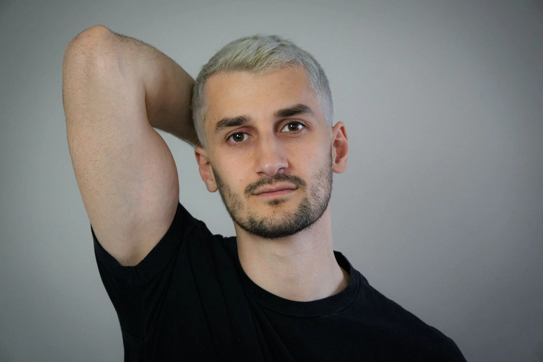 Junge Männerporträt mit grauen Haaren und Bart, trägt schwarzes T-Shirt, Blick in die Kamera, neutrale Haltung, grauer Hintergrund.