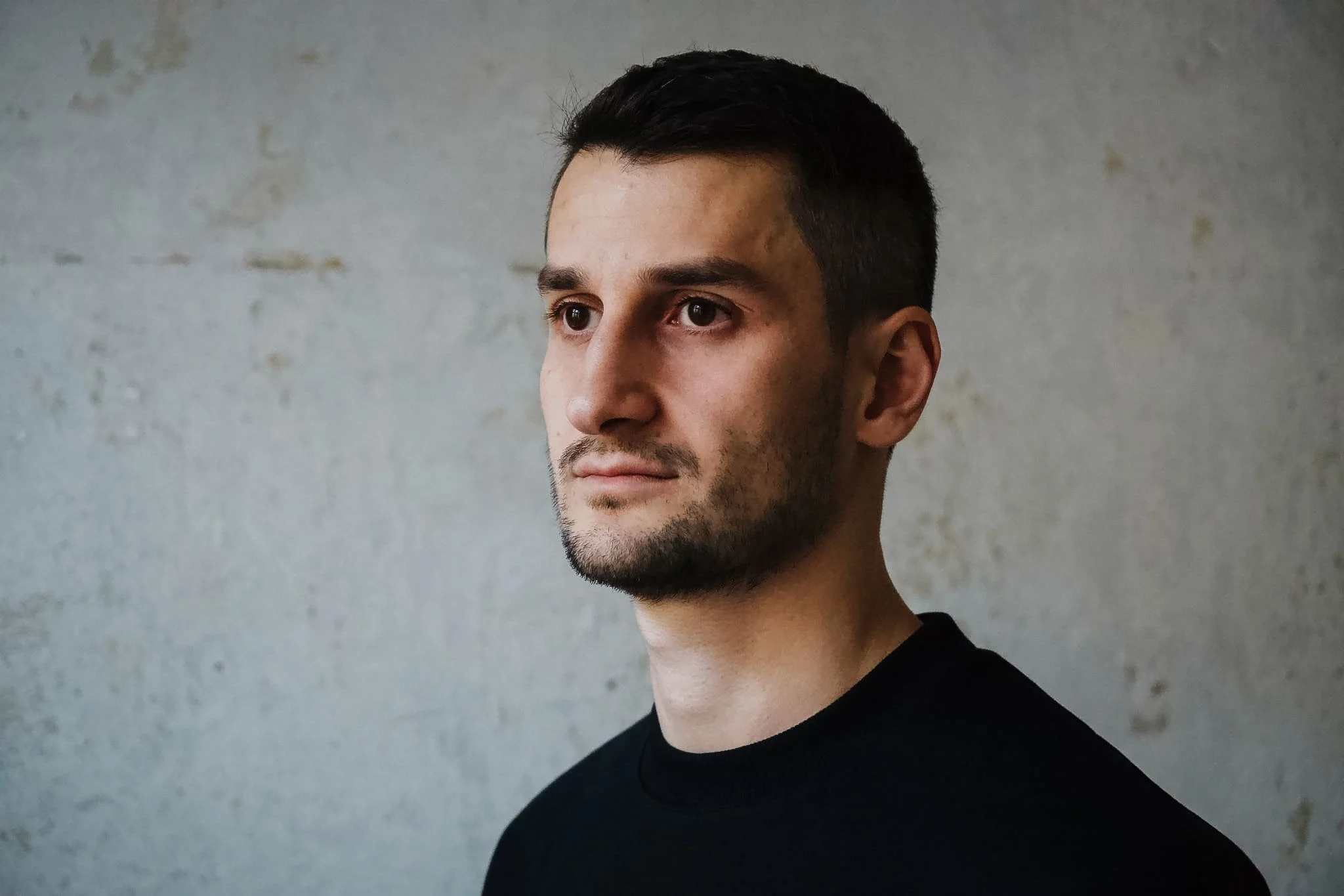 Ein junger Mann mit kurzen dunklen Haaren und hellbraunen Augen, trägt einen schwarzen Pullover, steht vor einer grauen Wand.