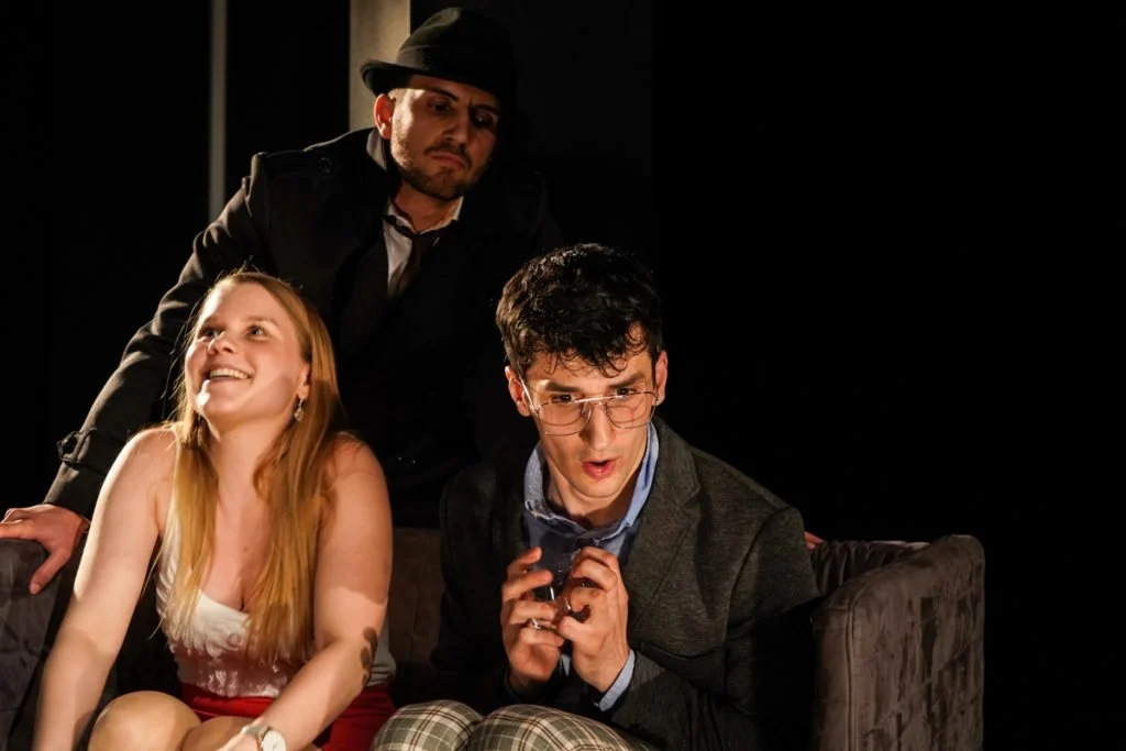 Drei Menschen auf einer Couch bei einer Theater- oder Filmszene, einer Frau lächelt, zwei Männer schauen interessiert.