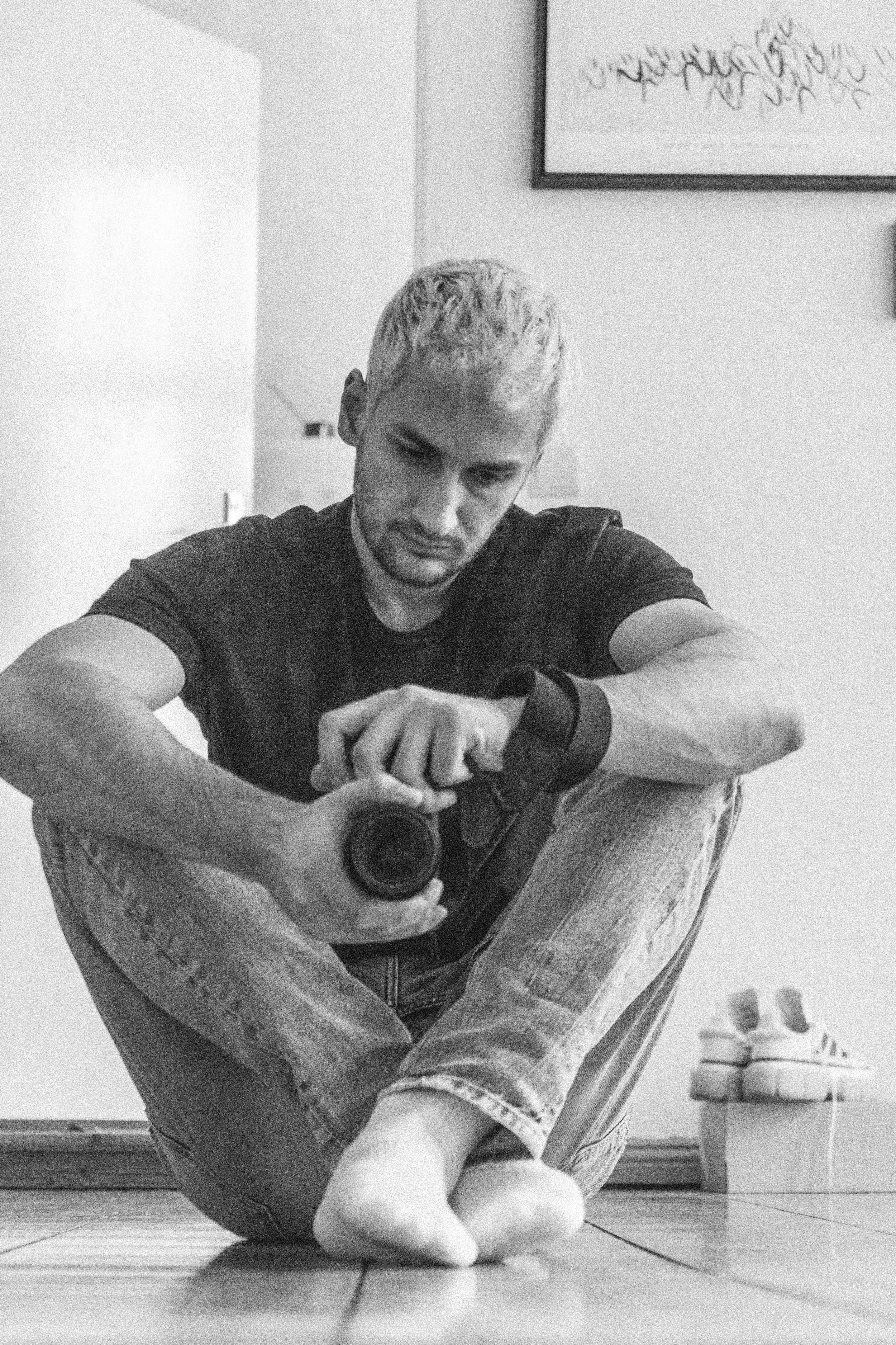 Junger Mann sitzt barfuß auf dem Boden, schaut auf seine Armbanduhr und fotografiert sich selbst mit einer Kamera. Im Hintergrund sind Schuhe in einem Korb und ein Wandbild zu sehen.