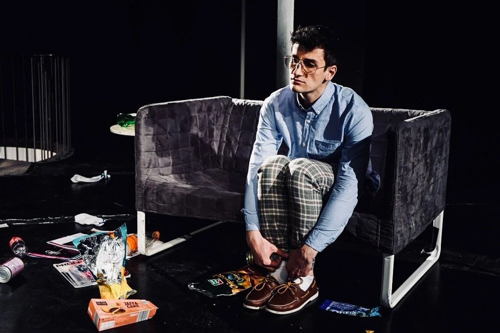 Ein junger Mann sitzt auf einem dunklen Sofa, zieht seine braunen Schuhe an und sieht nach unten. Er trägt eine hellblaue Hemd und karierte Hosen. Der Raum ist unordentlich mit Müll und Papier auf dem Boden. Die Umgebung ist dunkel, und es gibt eine schwarze Wand im Hintergrund.