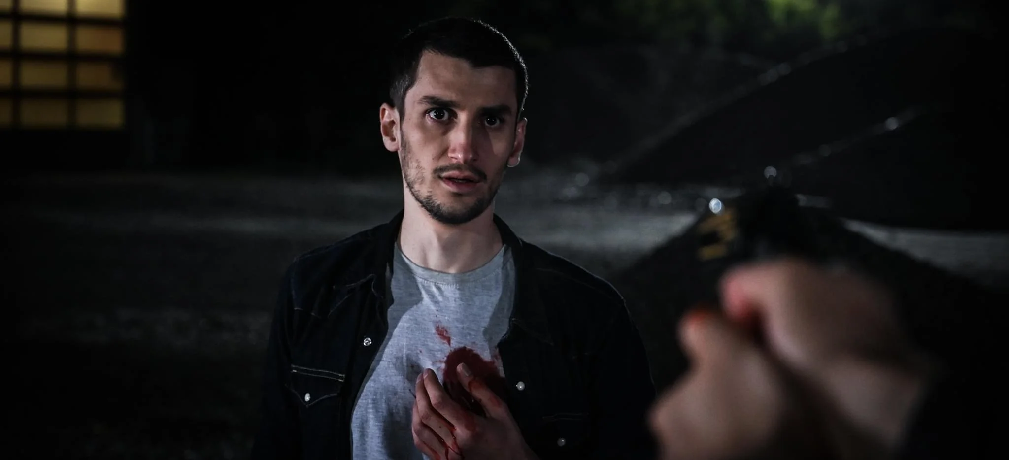 Ein Mann mit blutverschmiertem Shirt hält seine Hand an seine Brust, während im Vordergrund eine Waffe auf ihn gerichtet ist. Es ist Nacht draußen.