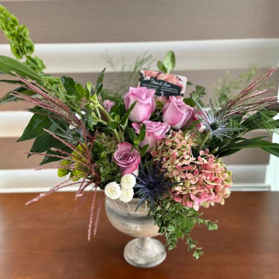A flower arrangement in a silver vase, featuring pink roses, hydrangeas, white button flowers, greenery, and decorative thistle-like blooms.