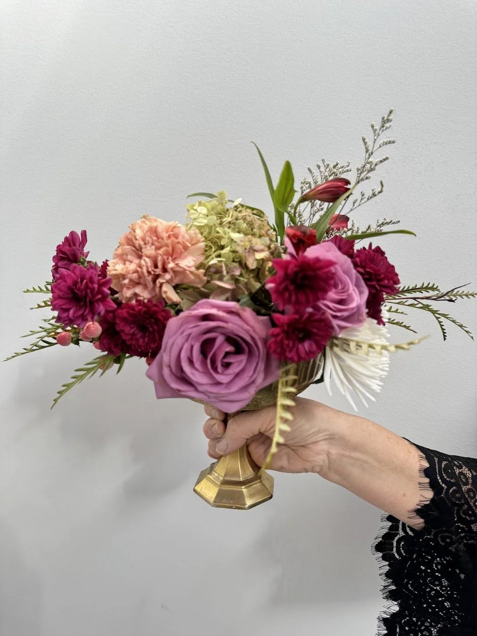 A hand holding a small bouquet of pink, purple, and red flowers with green foliage in a gold vase against a plain white background.